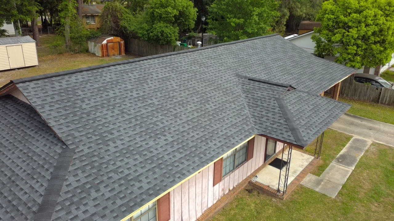 An aerial view of a house with a gray shingled roof surrounded by green trees and a fenced backyard. The house has a small concrete porch at the front.