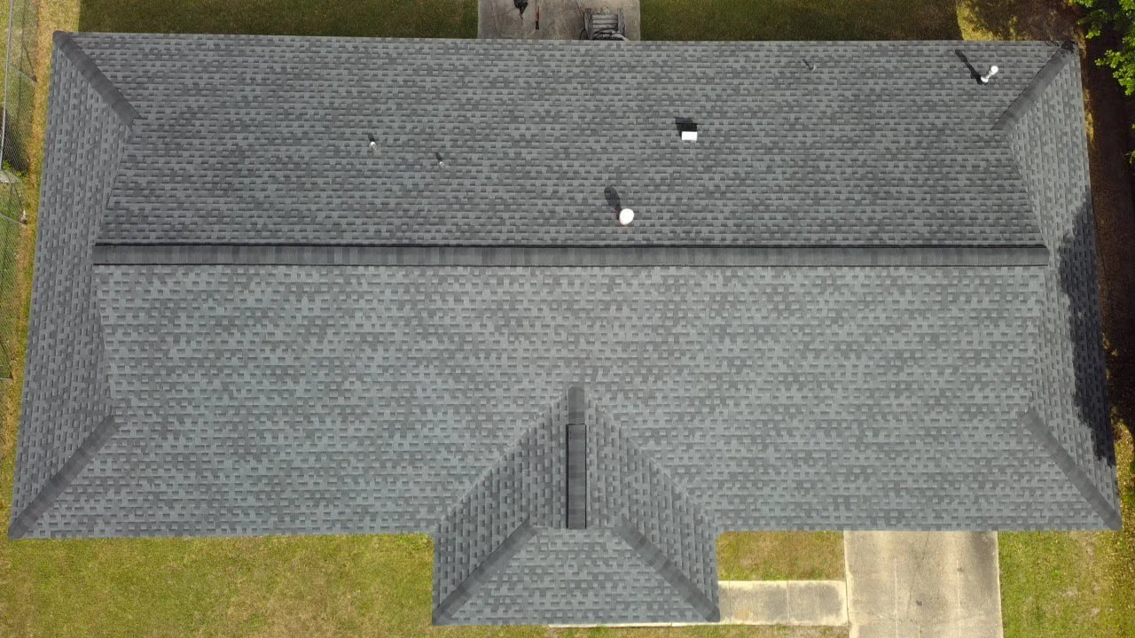 Aerial view of a gray shingle roof with multiple vents and a chimney, surrounded by green grass and concrete walkway.