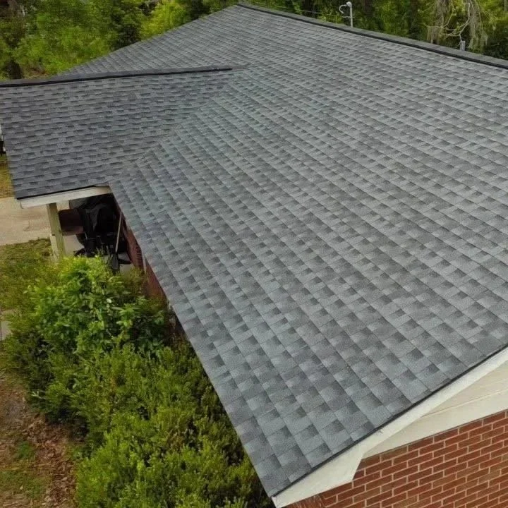 Aerial view of a house roof covered with gray asphalt shingles surrounded by green trees and bushes.