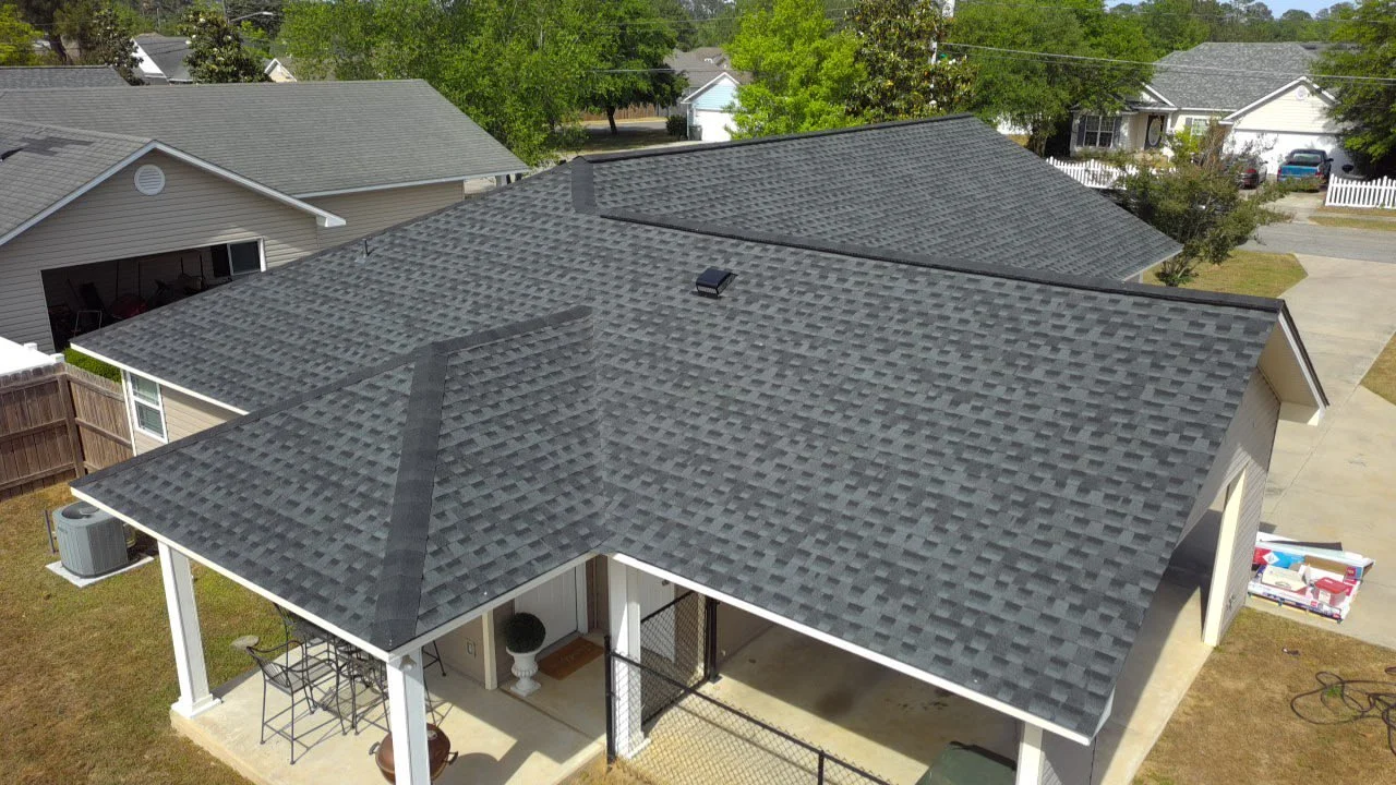 Aerial view of a house with gray asphalt shingle roof, white exterior walls, porch with outdoor furniture, and surrounding green yard with neighboring houses and trees.