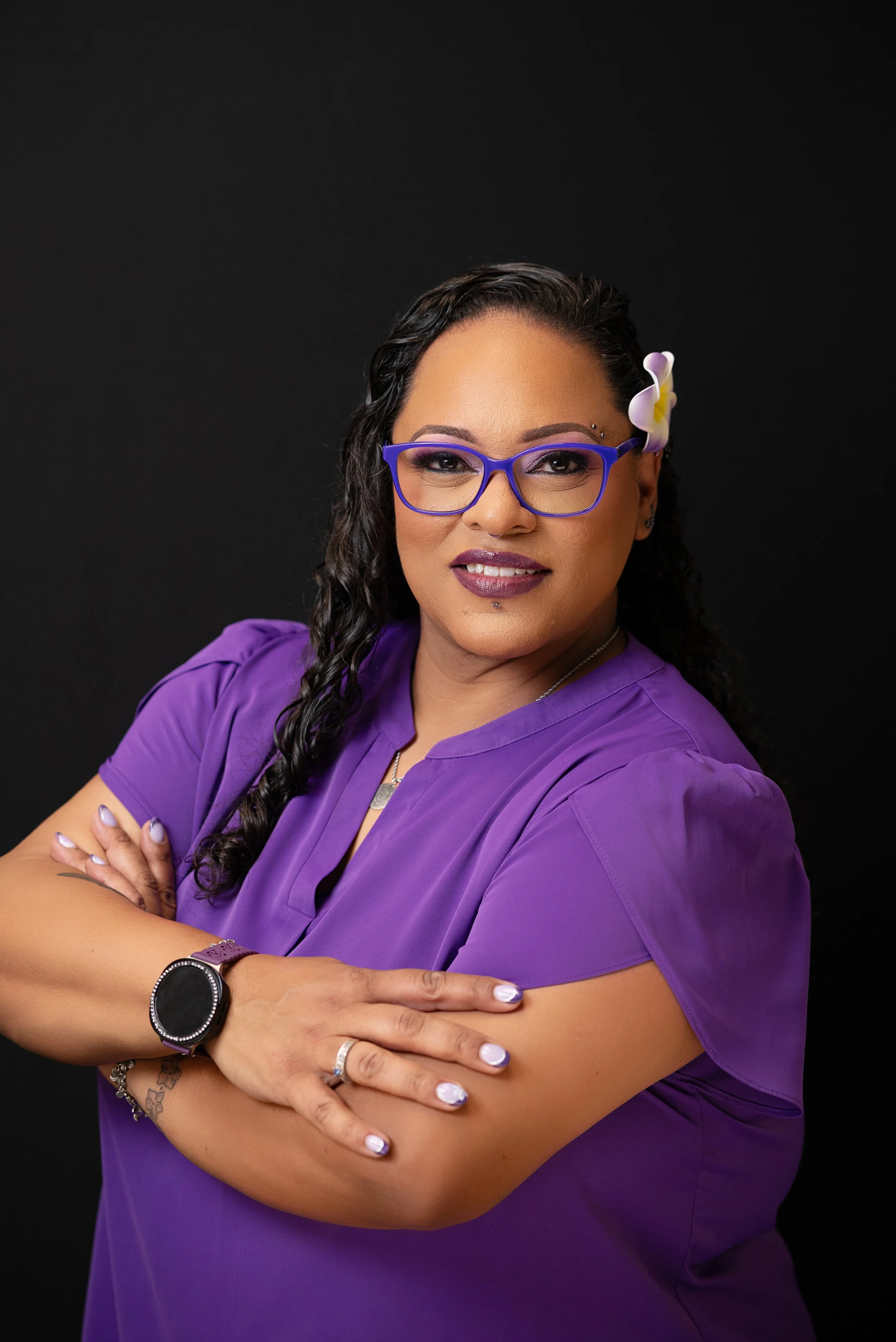 Woman with curly black hair, wearing purple glasses and a purple top, posed with crossed arms, against a black background.