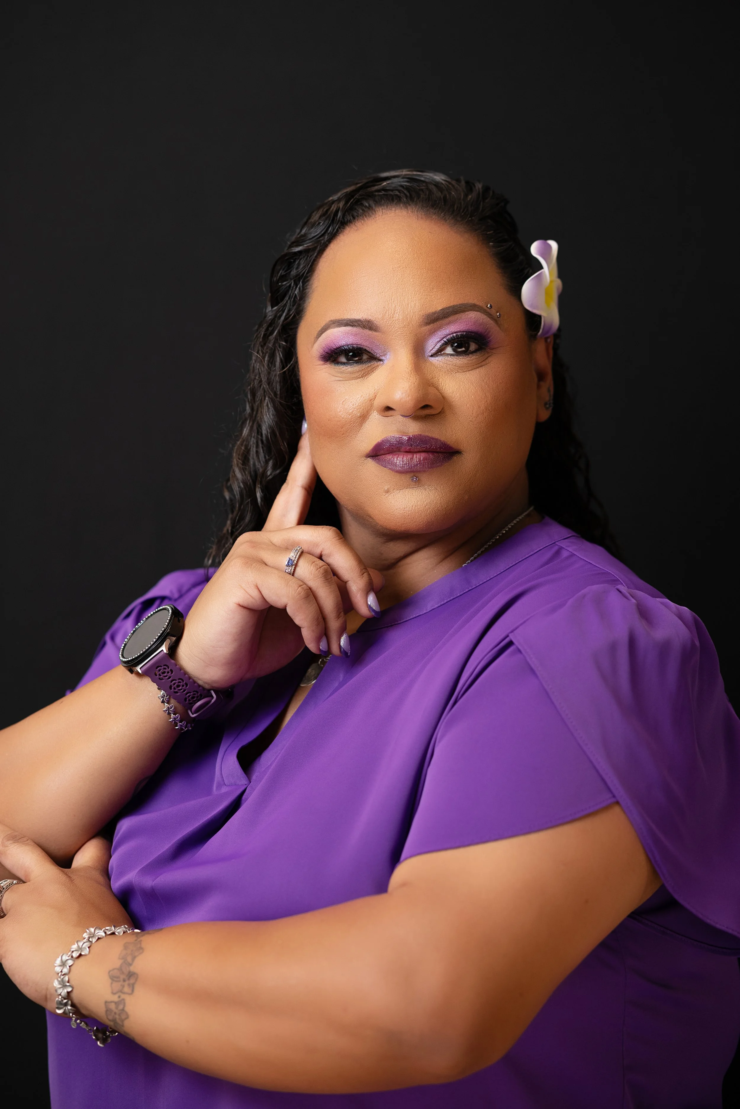 A woman with medium skin tone, curly dark hair, purple eyeshadow, purple lipstick, and a purple top, with a flower in her hair, jewelry, and tattoos, posing against a black background.
