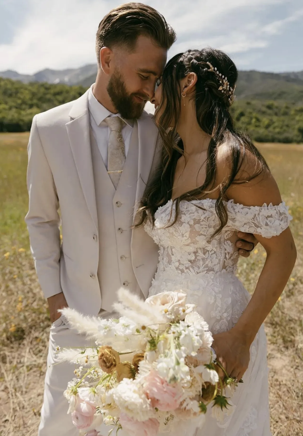 A newlywed couple in wedding attire sharing an intimate moment outdoors, with mountains and a partly cloudy sky in the background, and holding a bouquet of flowers.