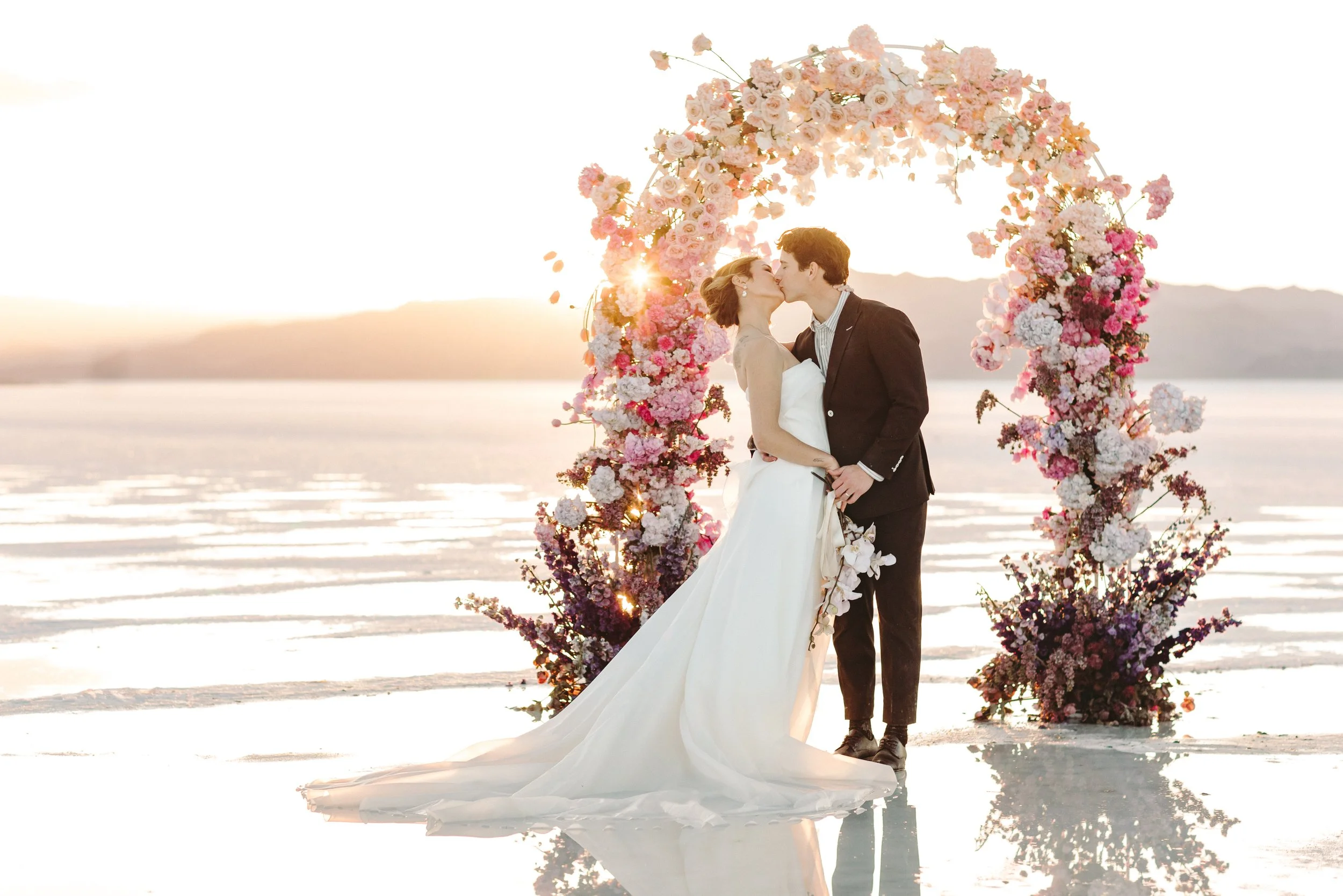 A newlywed couple kissing under a floral arch on a snowy landscape at sunset.