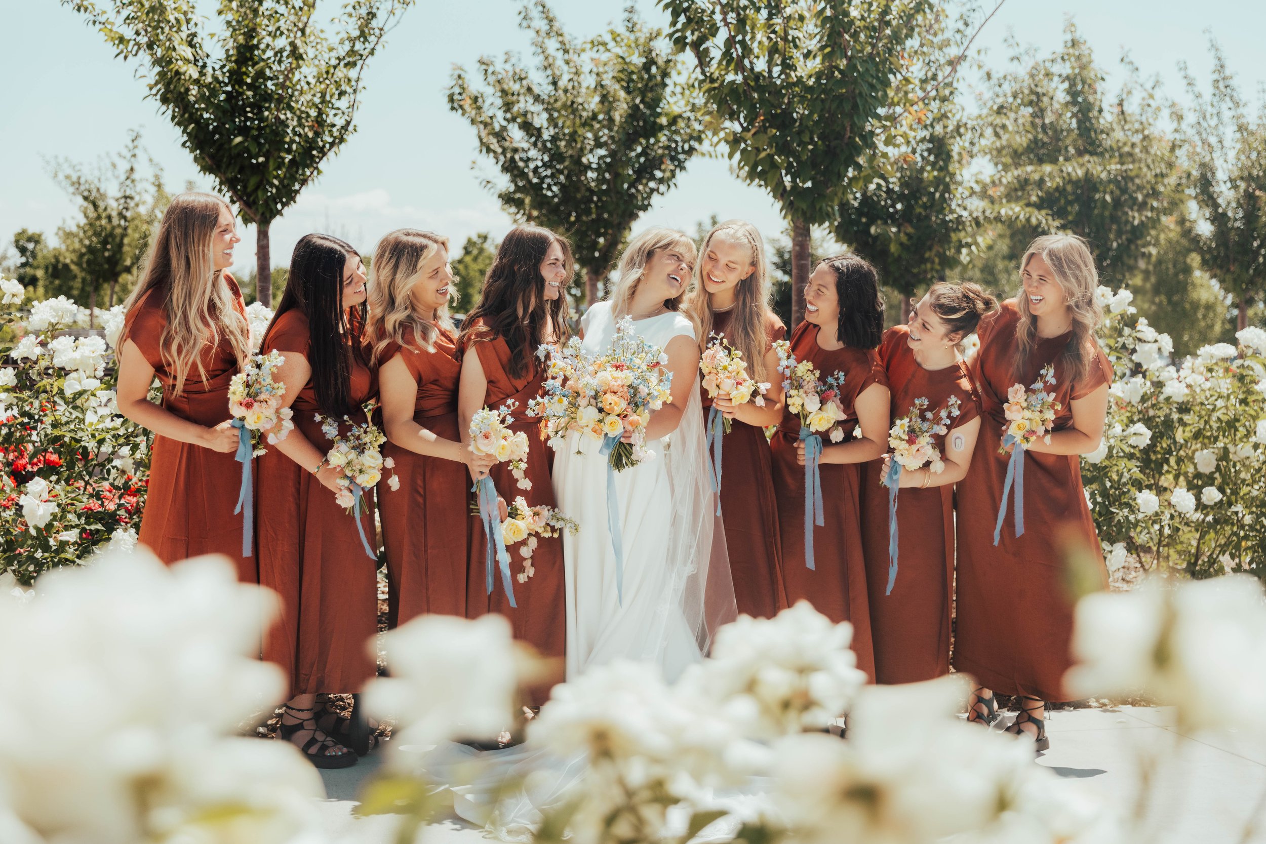 A group of women in matching brown dresses smiling and holding flower bouquets at an outdoor wedding ceremony, with trees and a clear sky in the background.