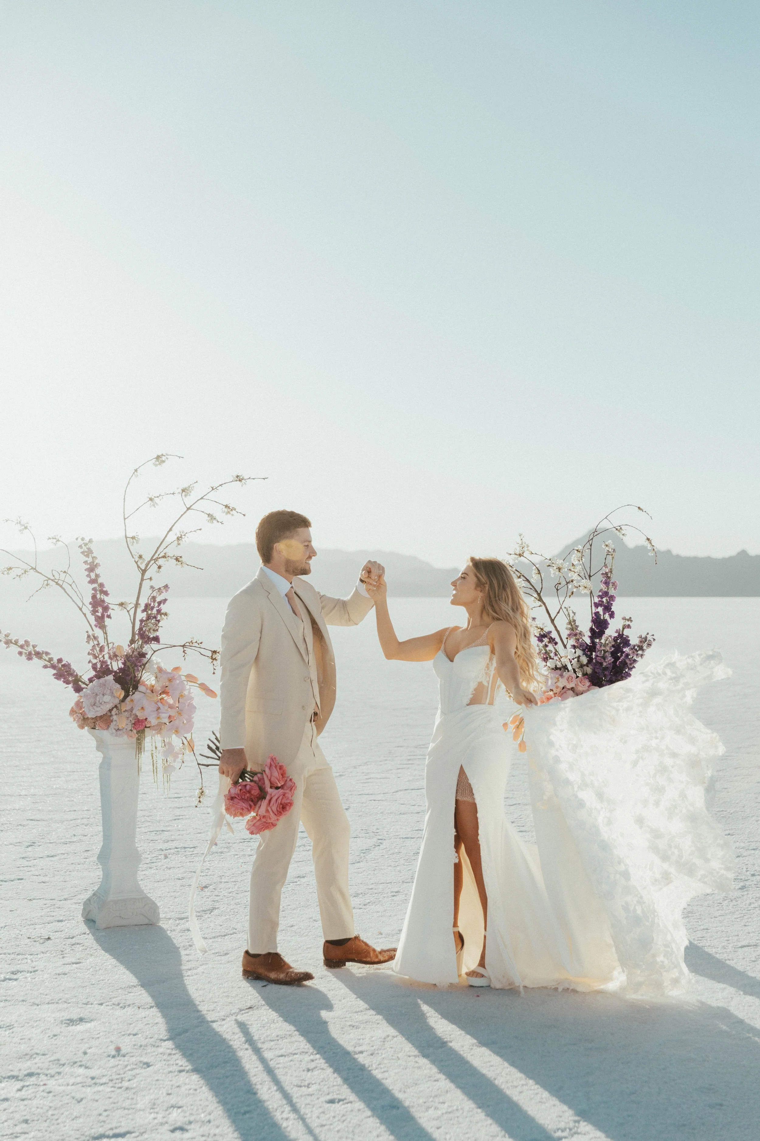 A couple getting married on a salt flat, with a man wearing a light-colored suit and a woman in a white wedding dress. They are holding hands and smiling at each other, surrounded by floral arrangements in white vases.
