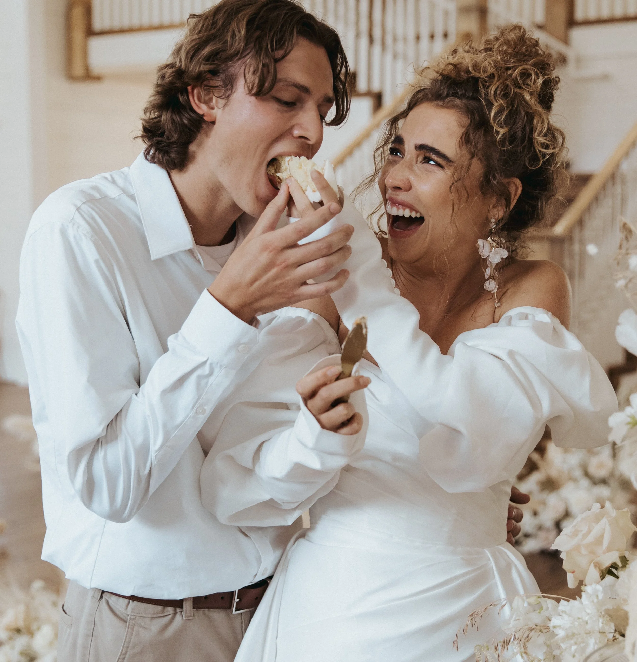 Two people, a young man and a woman, are joyfully sharing a piece of cake, with both smiling and looking at each other. They are dressed in white clothing, and the background appears to be indoors with a staircase and floral decorations.