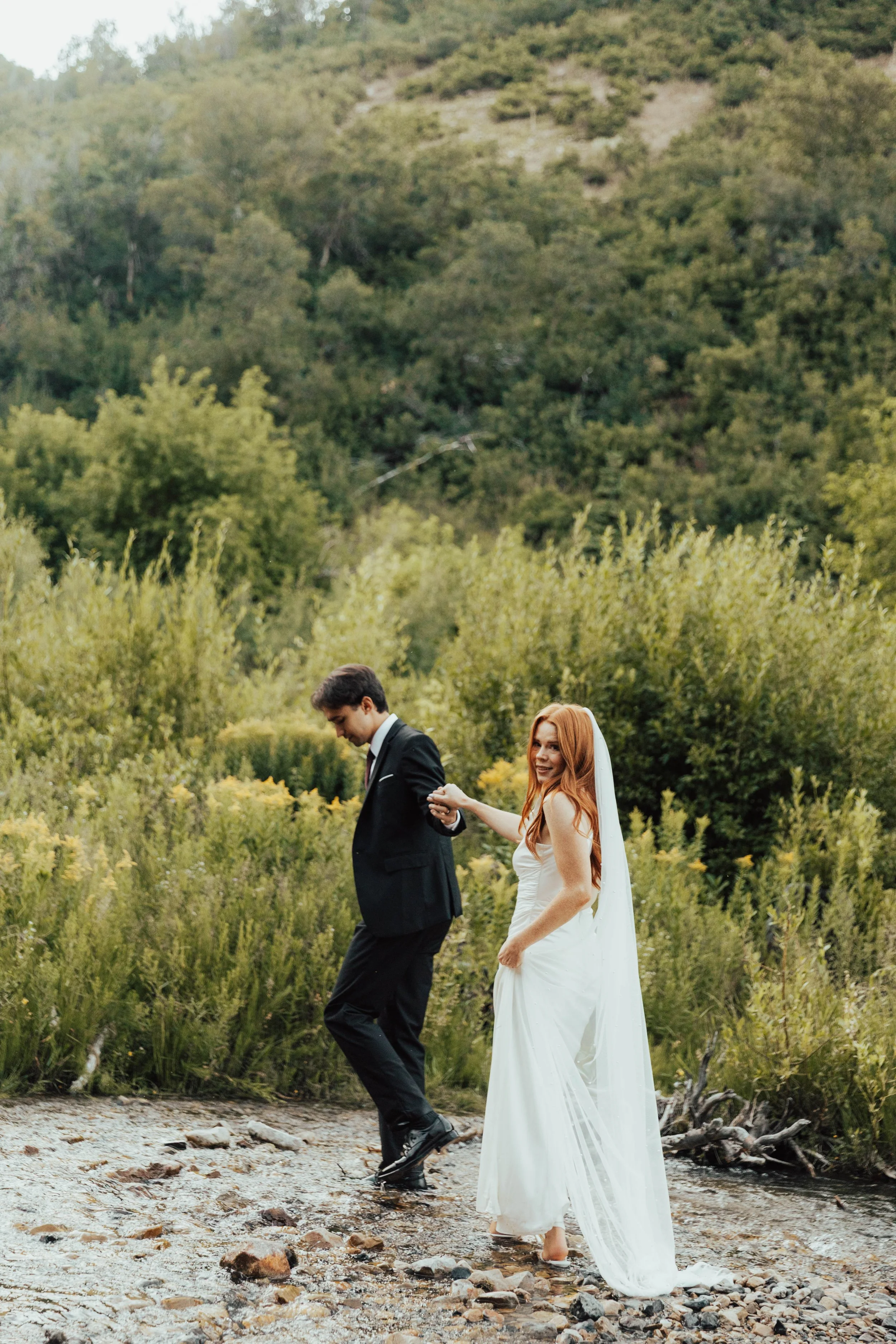 A bride and groom holding hands and walking through a shallow stream in a lush green forested area with hills in the background during daytime.