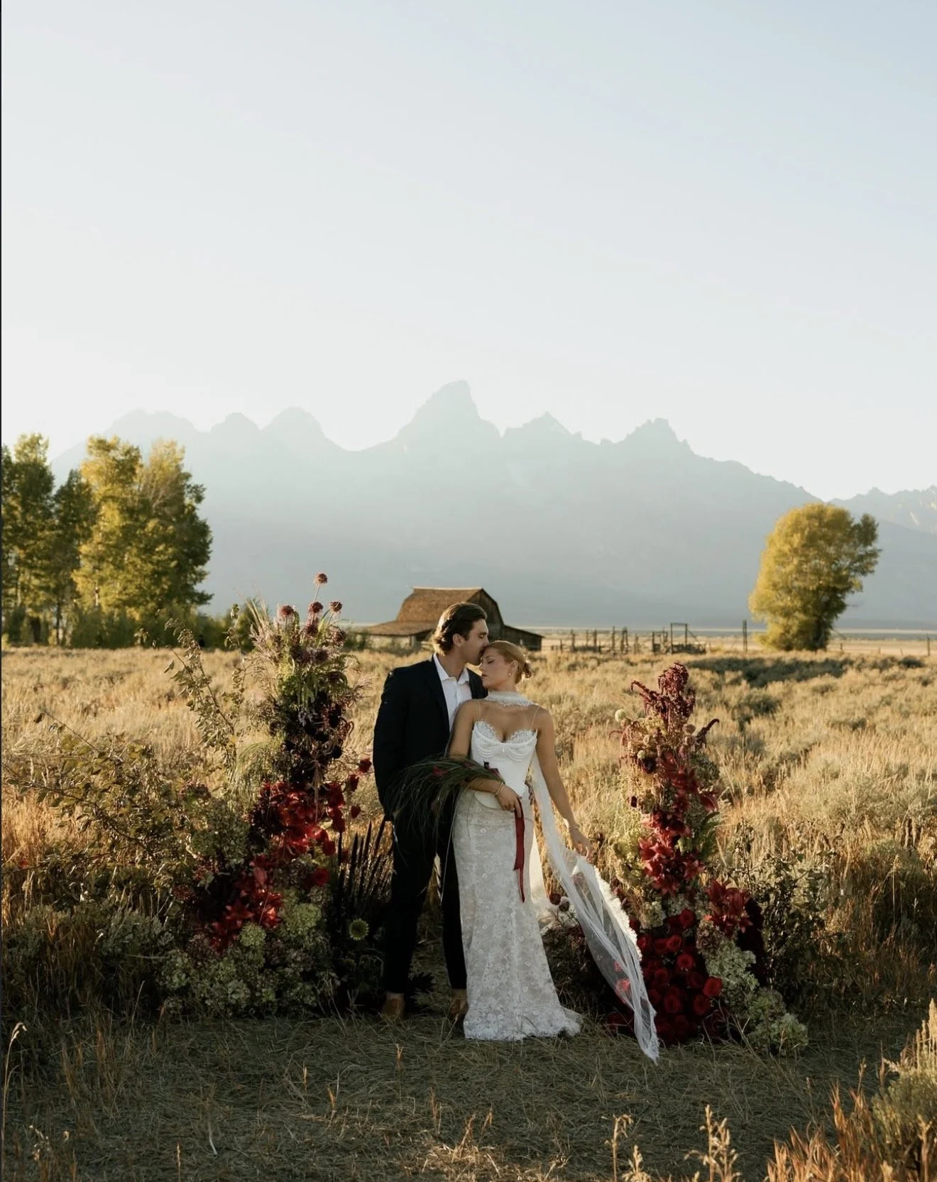 A bride and groom share a romantic moment in a field during sunset, surrounded by floral arrangements, with mountains in the background.
