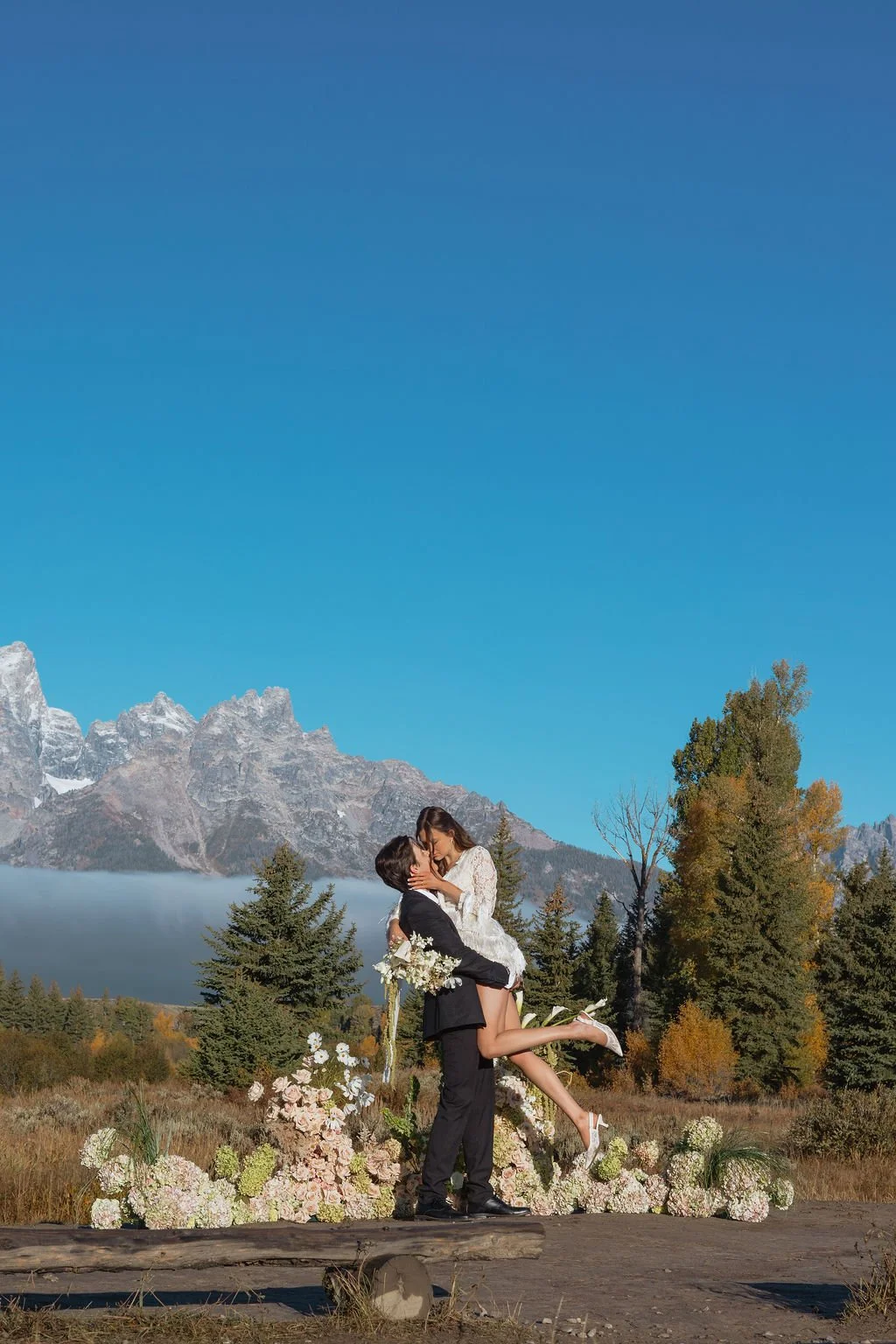 A couple in wedding attire sharing a kiss outdoors, with a mountain landscape and clear blue sky in the background, surrounded by flowers and greenery.