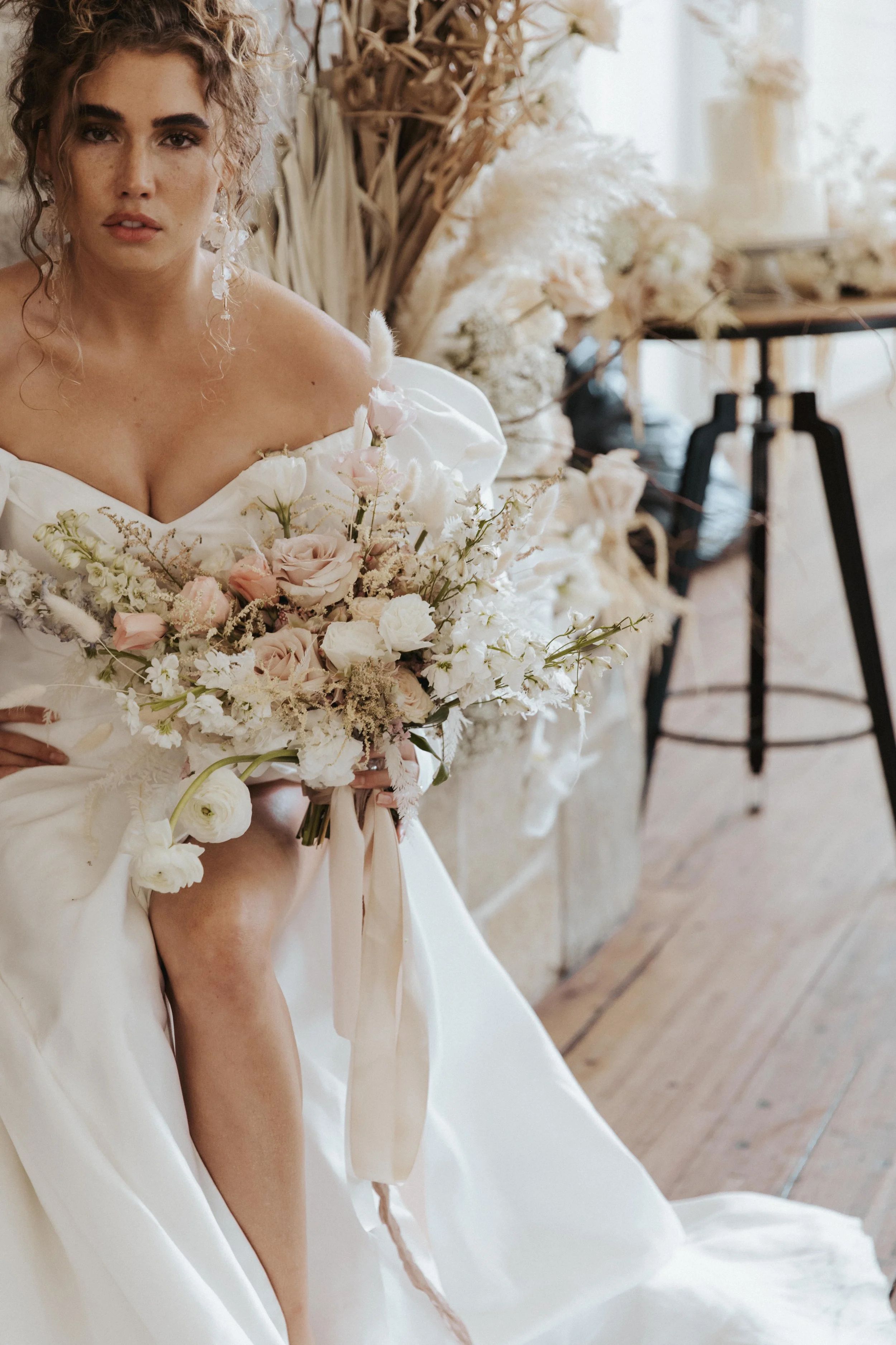 A woman in a bridal gown holding a bouquet of pale pink and white flowers, standing in a room decorated with dried flowers and a wedding cake in the background.