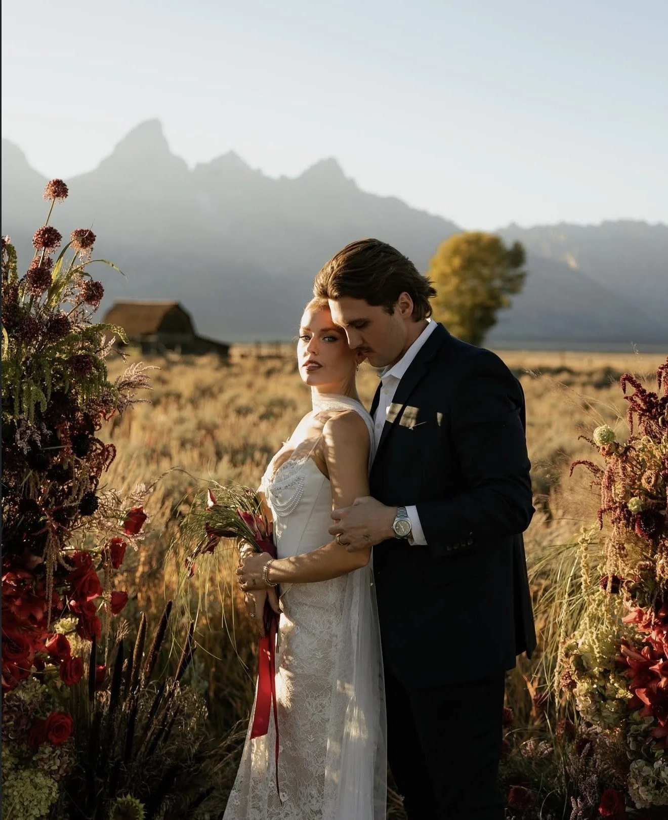 A bride and groom standing closely together in a field of flowers at sunset with mountains in the background. The bride is wearing a white lace wedding dress and holding a bouquet, while the groom is in a black suit. They are embracing each other.
