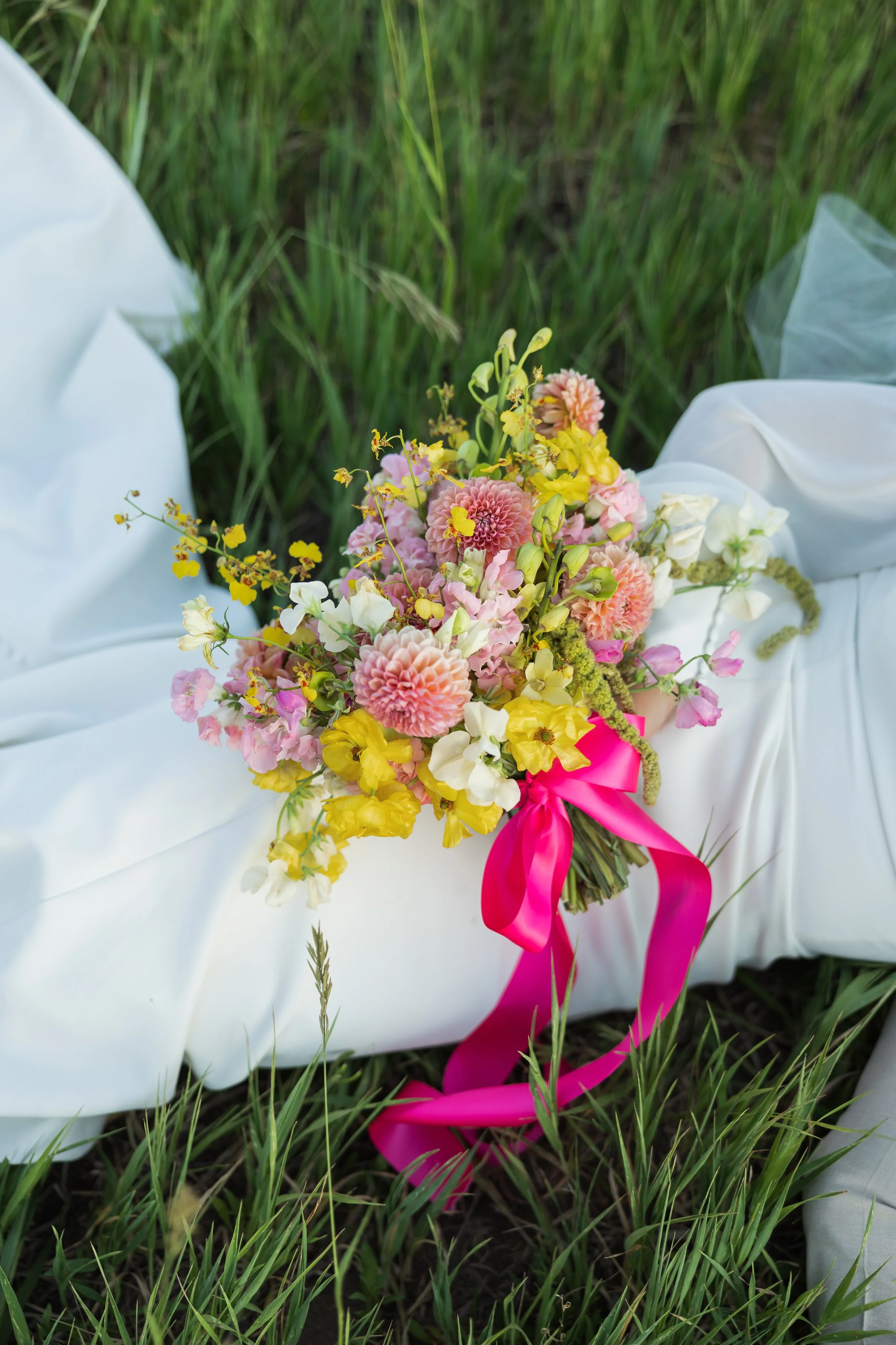 A bouquet of pink, white, and yellow flowers with green foliage and pink ribbon resting on white fabric outdoors on grass.