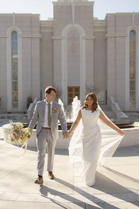 A bride and groom holding hands, walking in front of a large church with a fountain, the bride in a white wedding dress and the groom in a light gray suit, holding a bouquet of flowers.