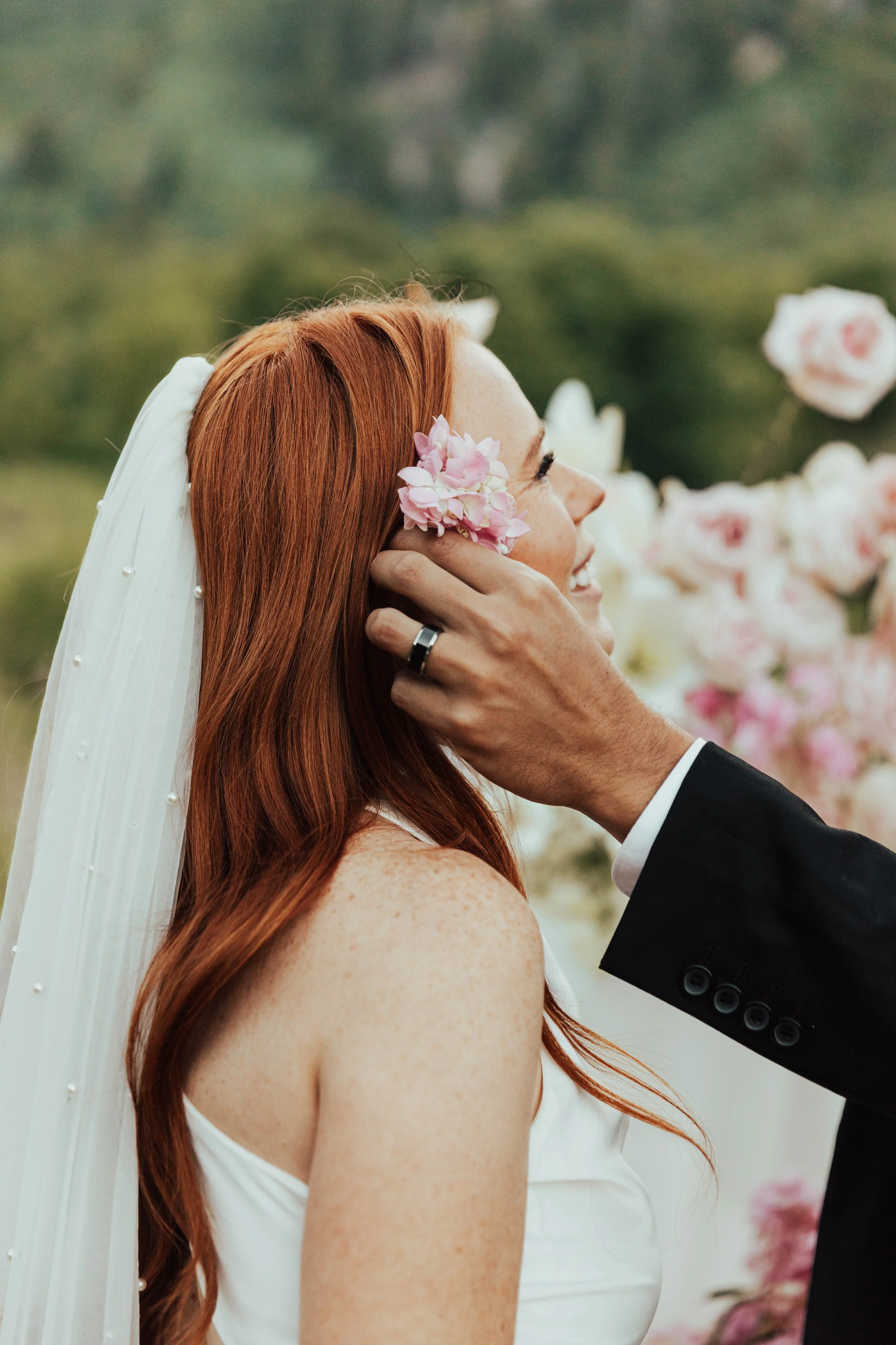 A red-haired woman in a white dress and veil, smiling as a man in a black suit touches her face. She has a pink flower tucked behind her ear and appears happy. Out-of-focus pink and white flowers are in the background.