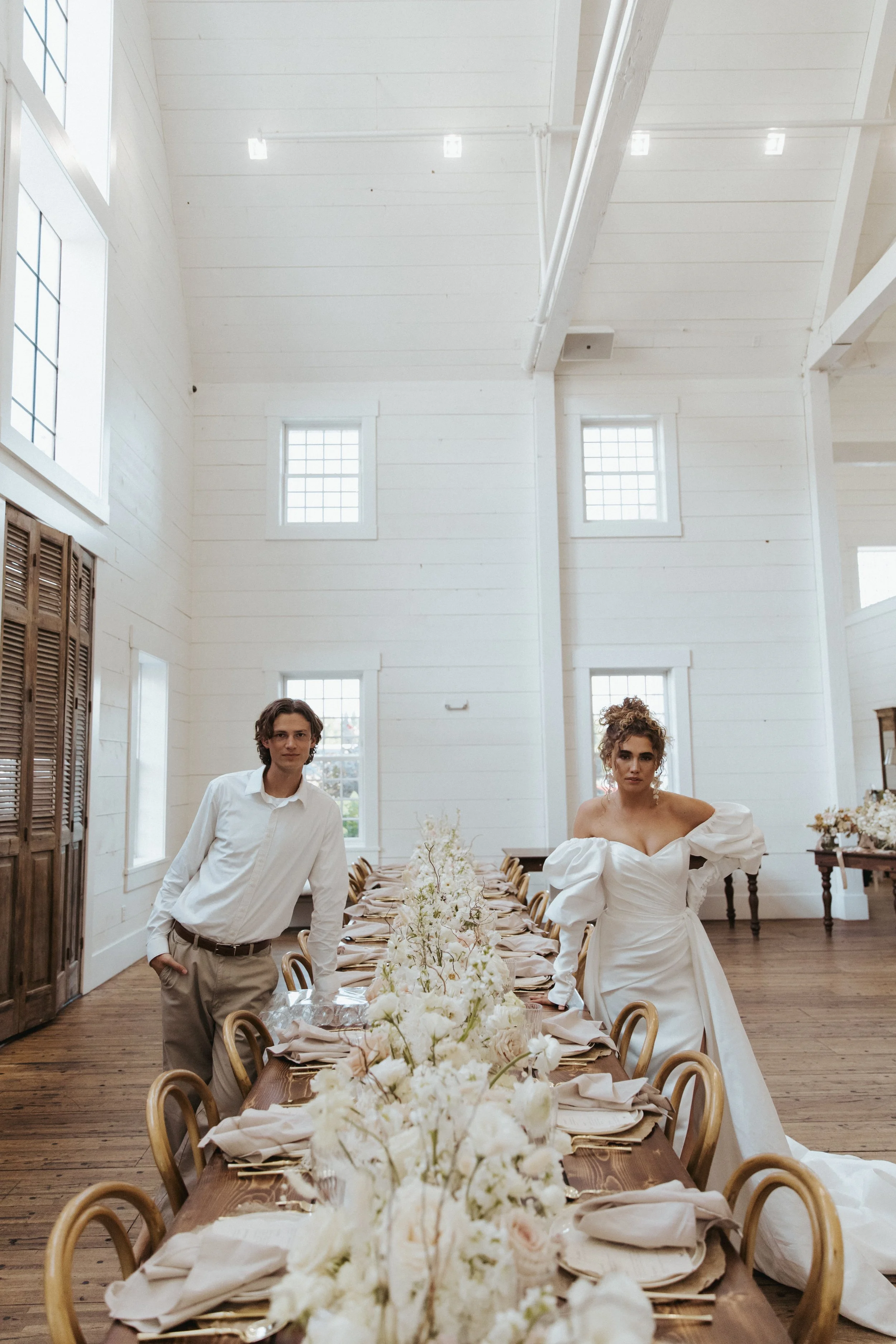A man and woman standing at a long dining table decorated with white flowers inside a bright room with high ceilings and large windows.