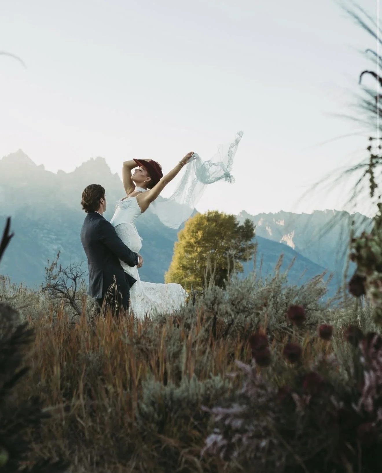A couple in wedding attire stands in a field with mountain landscape in the background. The groom holds the bride, who lifts her arms with a flowing veil, against a bright sky with trees and foliage around them.
