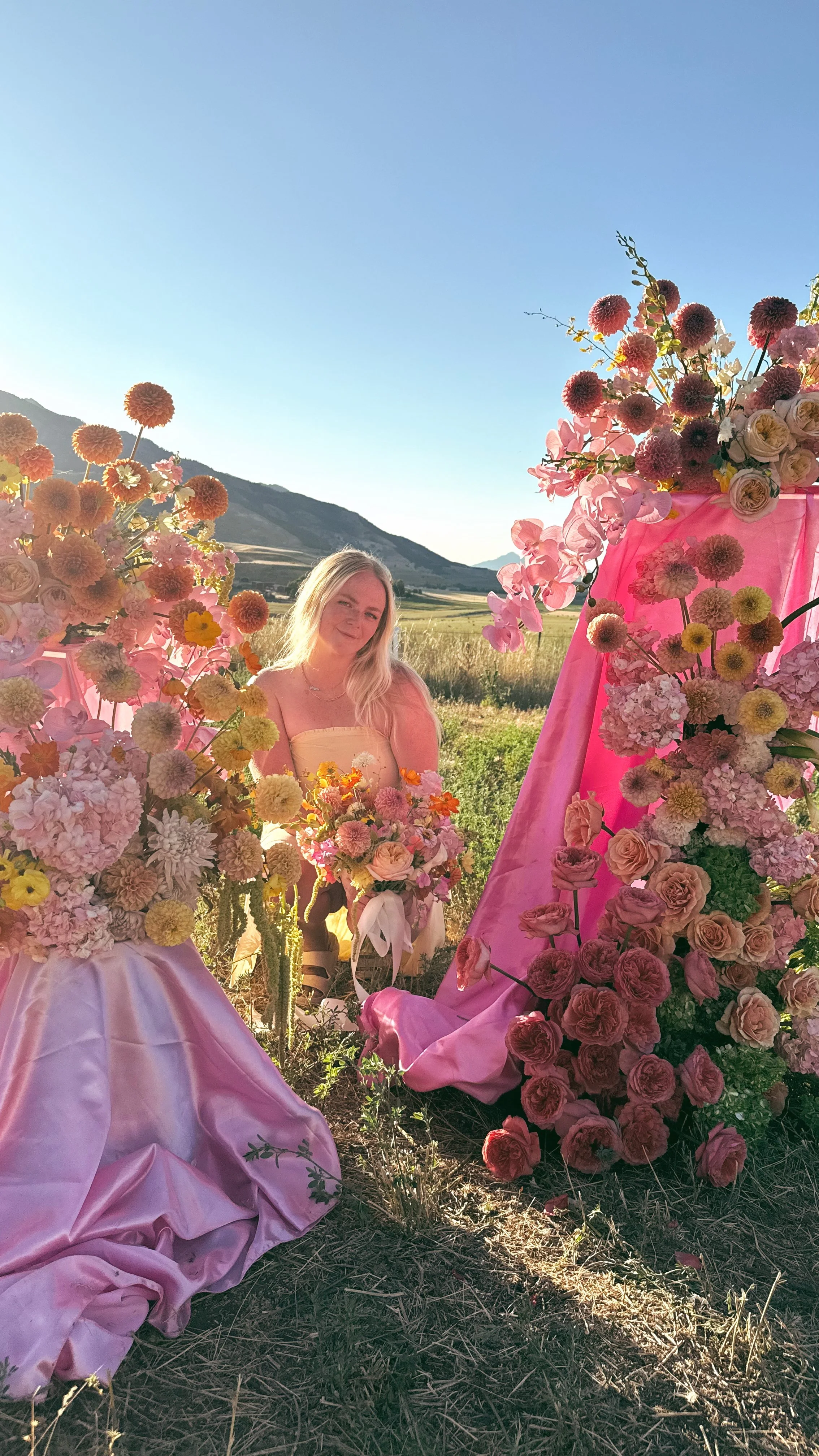 A woman with long blonde hair in a beige dress sitting among pink, peach, and white flowers arranged on pink fabric-draped structures outdoors in a field with mountains and a clear blue sky in the background.
