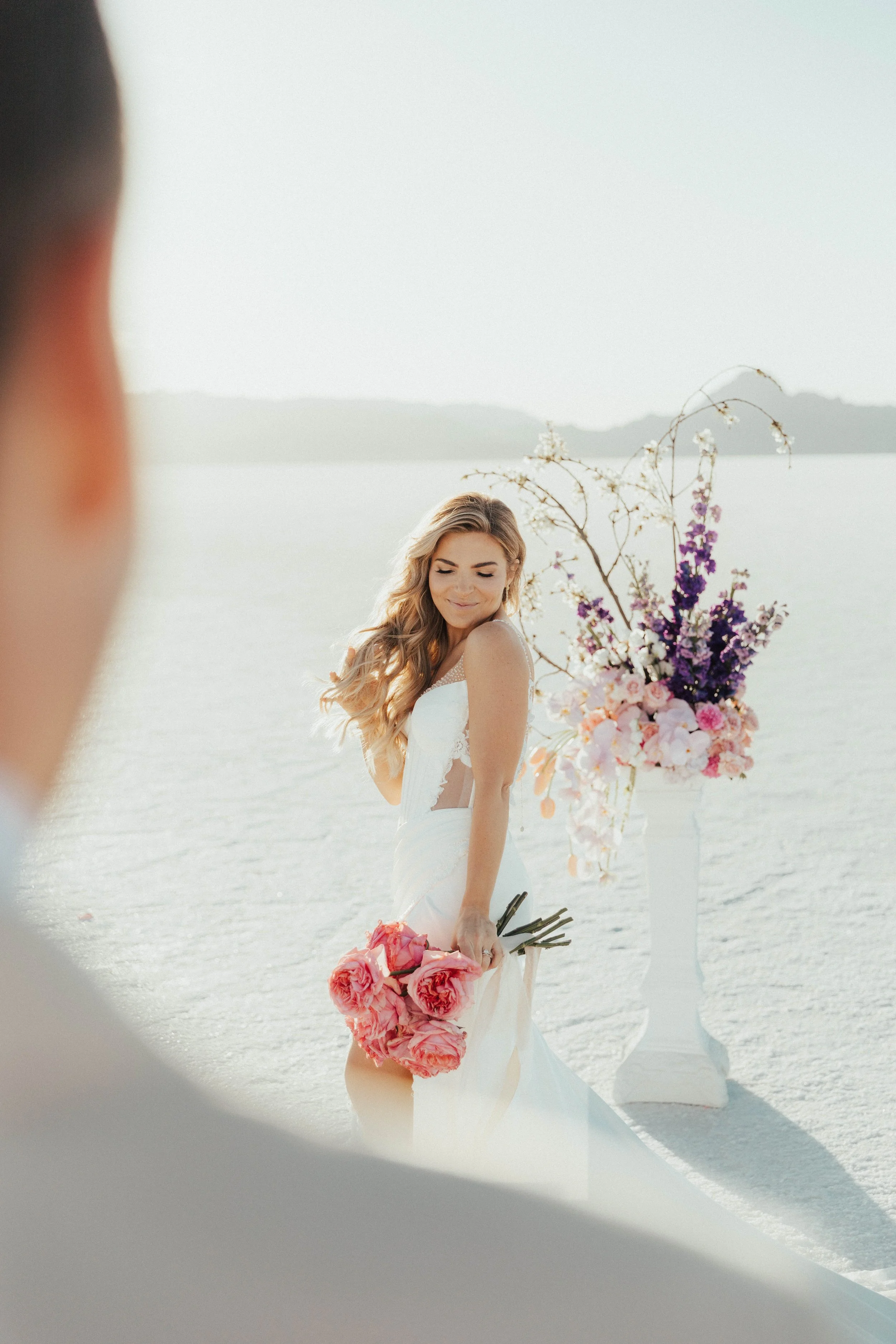 A bride with long, wavy blonde hair wears a white wedding dress and holds a bouquet of pink roses, standing outdoors near a large floral arrangement with pink, purple, and white flowers, with a body of water and mountains in the background.