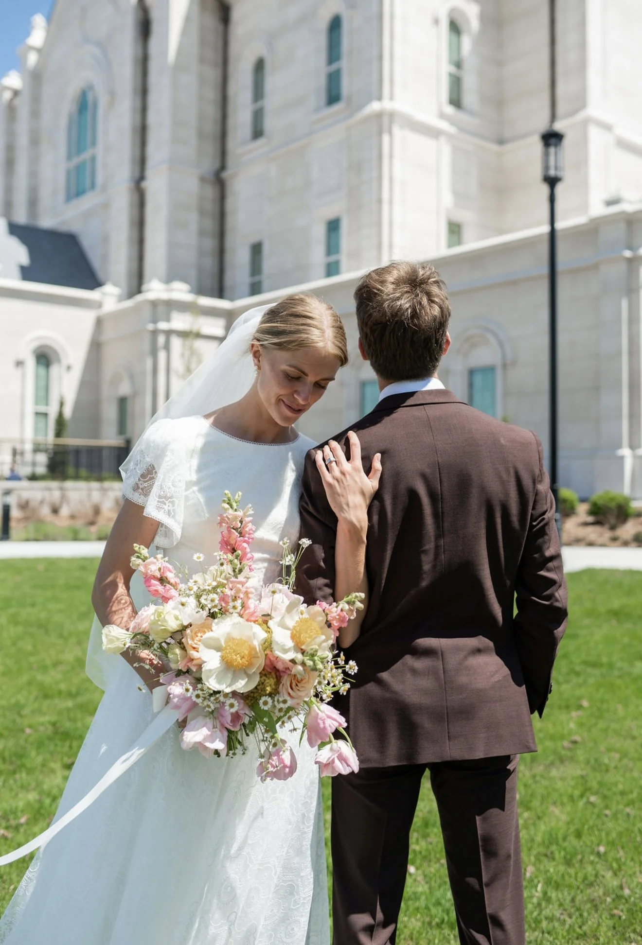 A bride and groom standing outdoors in front of a large stone building, sharing a moment during their wedding, with the bride holding a colorful bouquet of flowers.