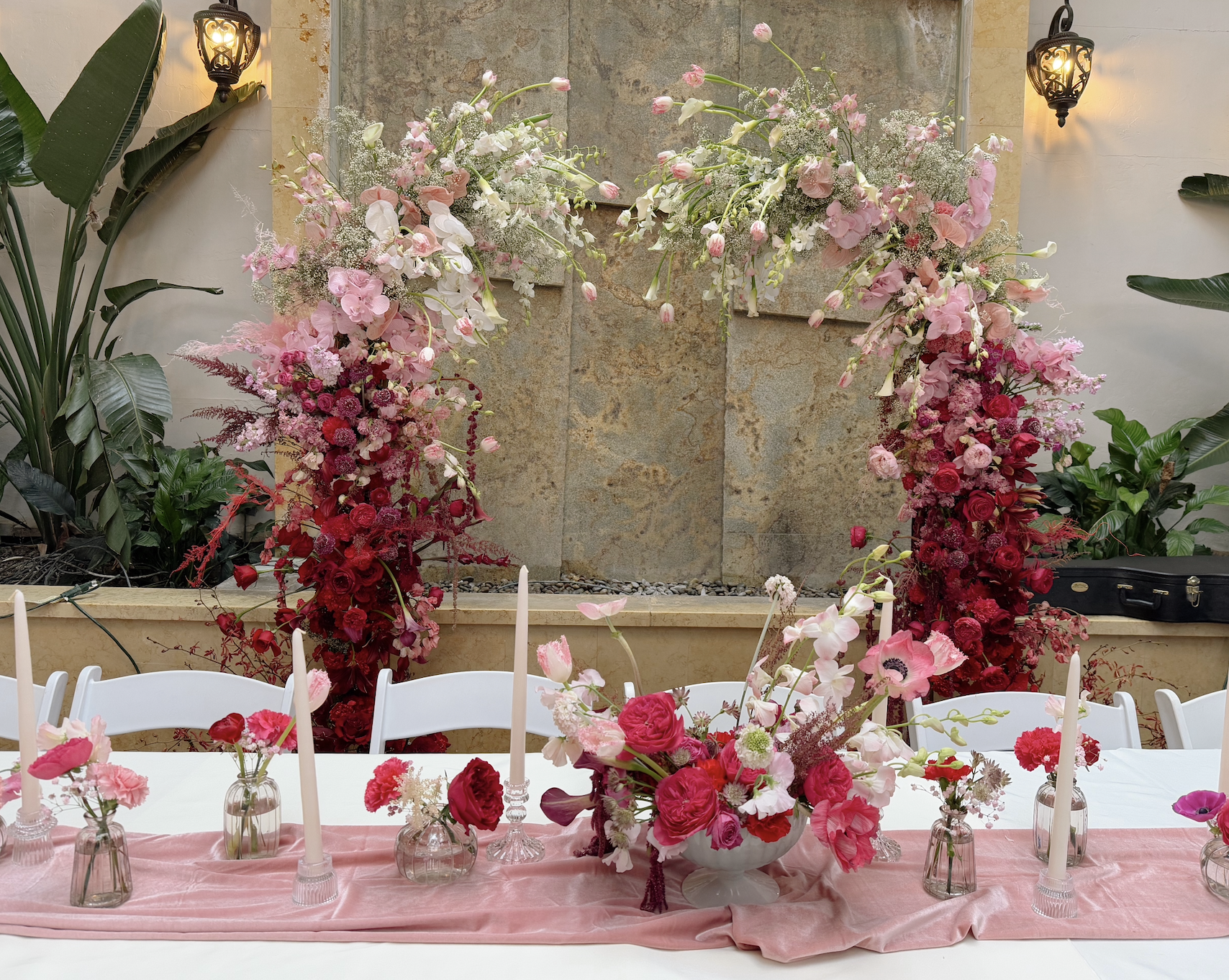A long banquet table decorated with pink, red, and white floral arrangements in glass vases, topped with tall white candles, set against a backdrop of a stone wall with greenery and decorative lanterns.
