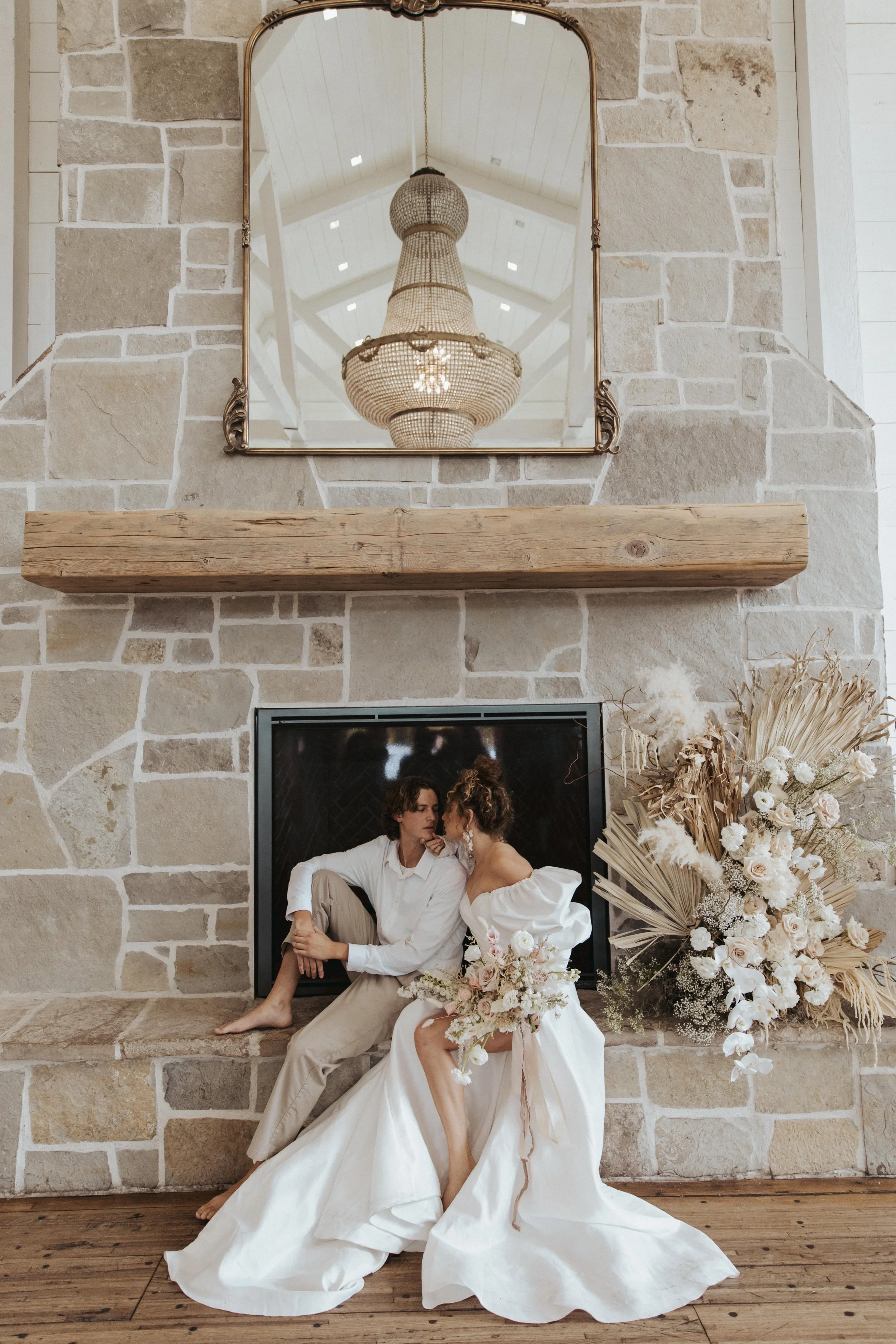 A couple dressed in elegant attire sitting in front of a stone fireplace, surrounded by a large bouquet of white and cream flowers, with a decorative mirror and chandelier overhead.