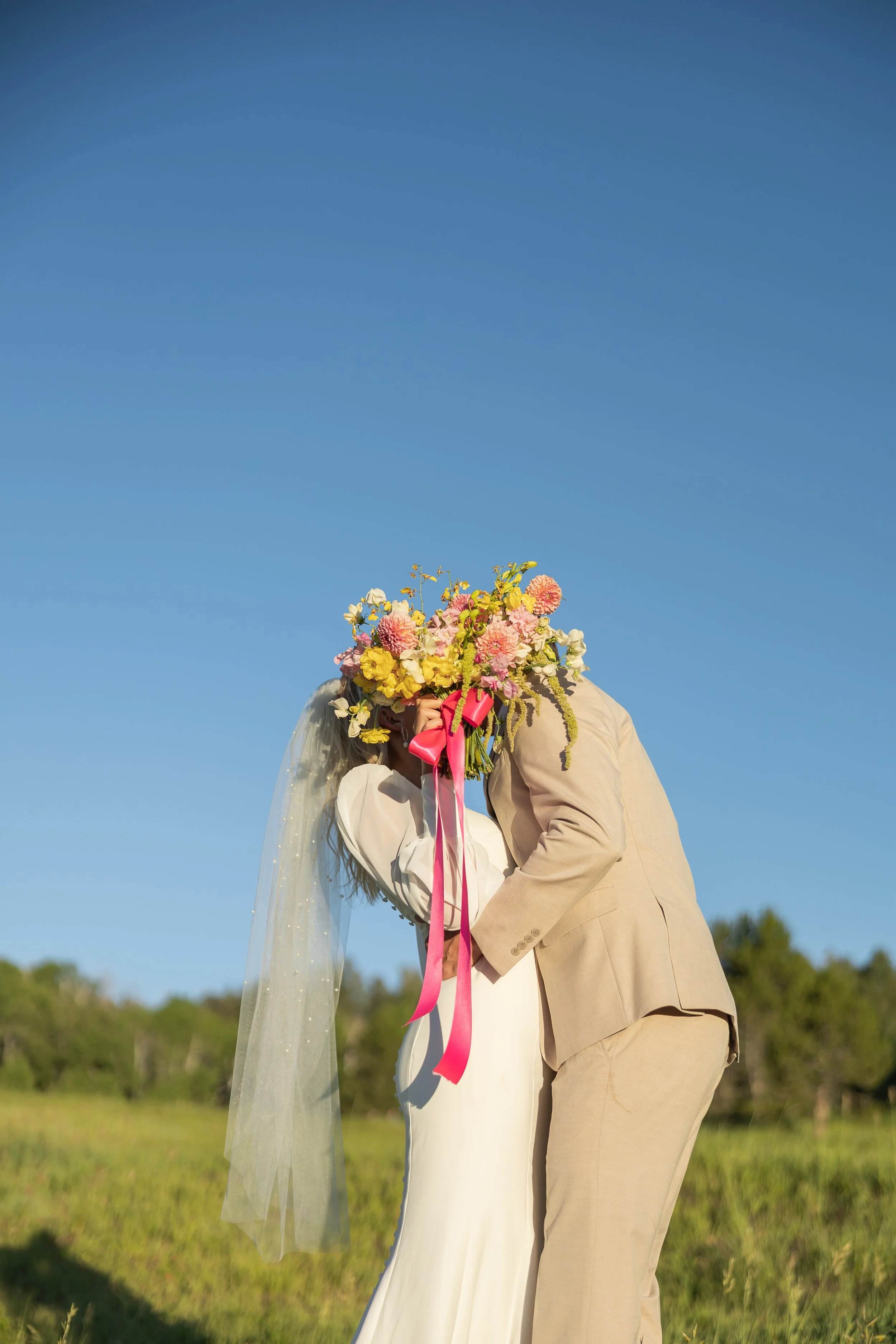 A bride and groom embracing outdoors, with the groom covering the bride's face with a bouquet of pink, yellow, and white flowers against a clear blue sky.