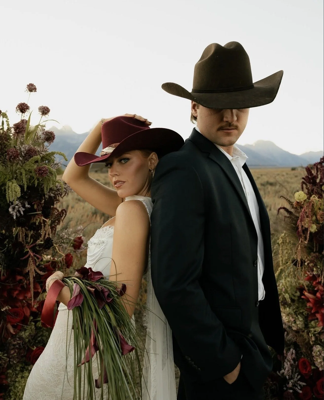 A man and a woman dressed in wedding attire standing back-to-back outdoors with mountains in the background, surrounded by flowers. The woman is holding a bouquet and tipping her hat, while the man is wearing a cowboy hat, with his hands in his pocke