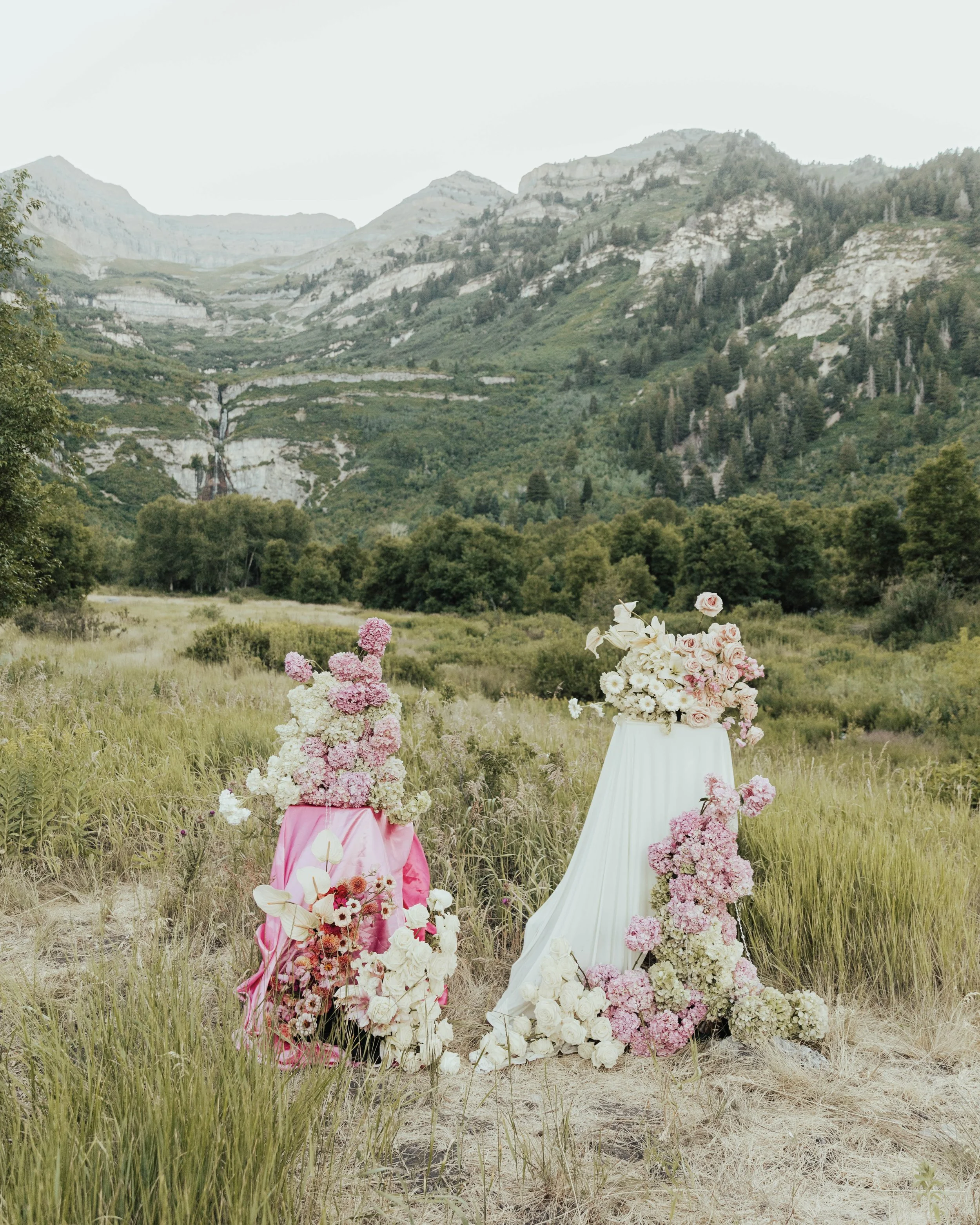 Field of green grass and wildflowers with two floral arrangements on stands, located in a scenic valley surrounded by mountains and forest.