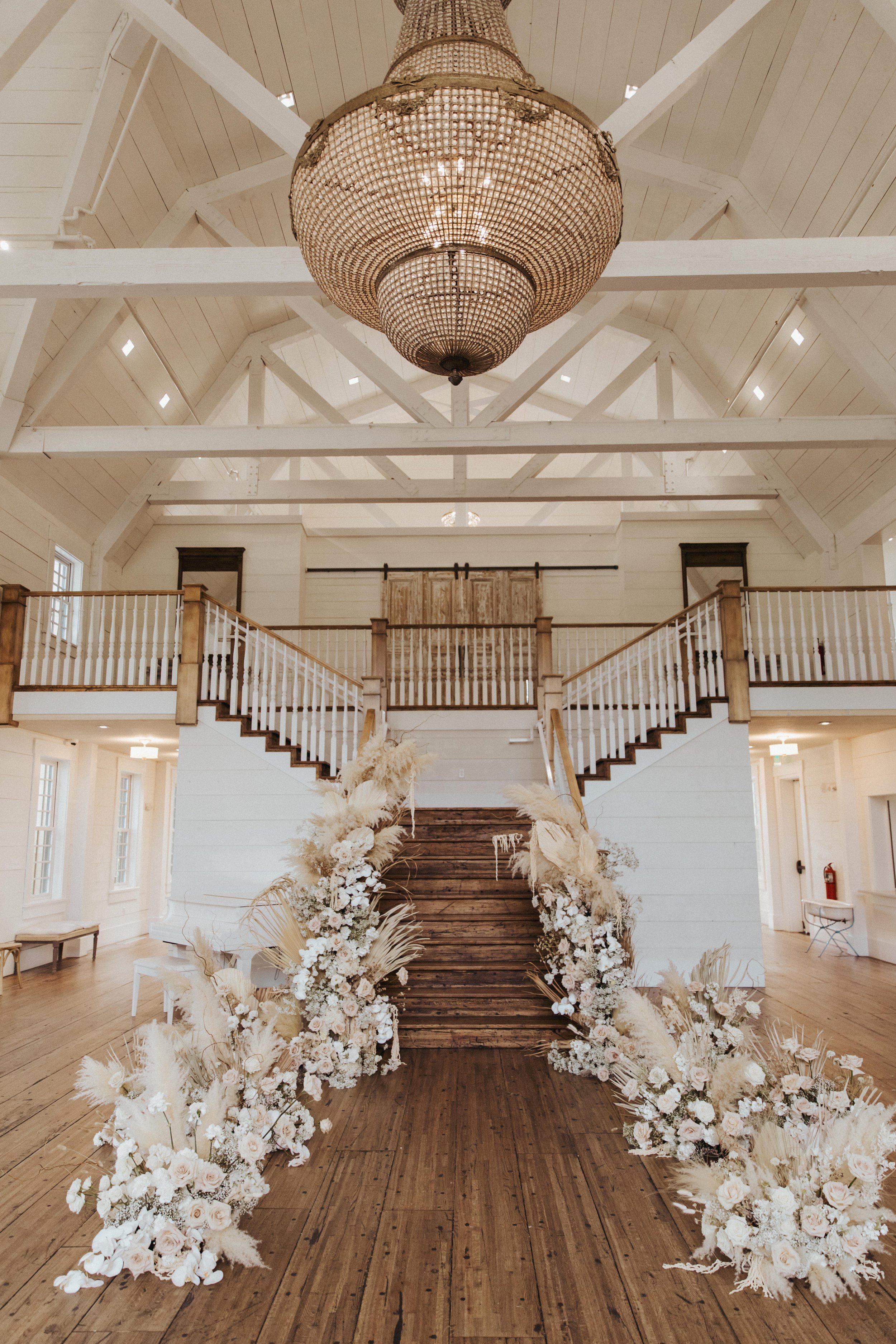A grand interior space with high vaulted ceiling, white exposed beams, a large ornate chandelier, a wooden staircase decorated with white and neutral-toned floral arrangements and pampas grass, leading to a balcony with a wooden railing.