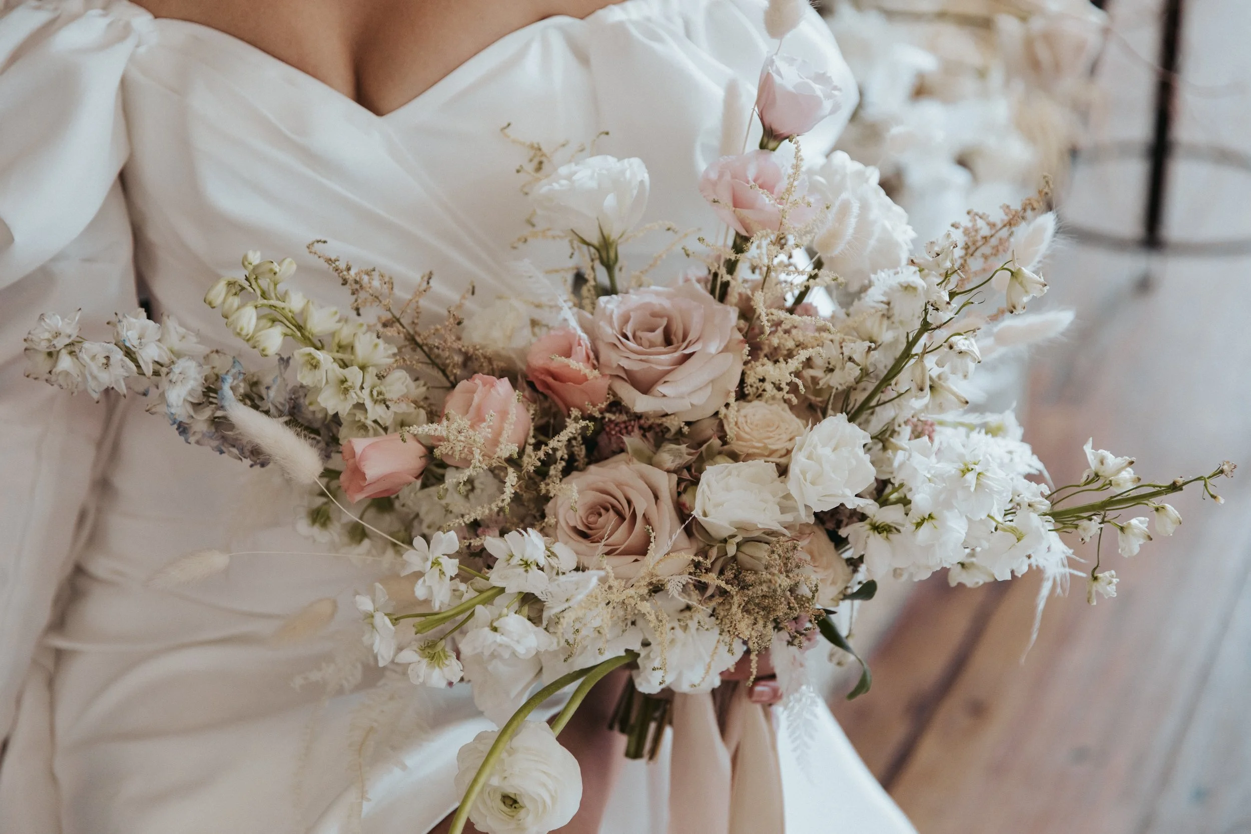 A person holding a bridal bouquet with various pale-colored flowers including roses and greenery, dressed in an ivory wedding gown.