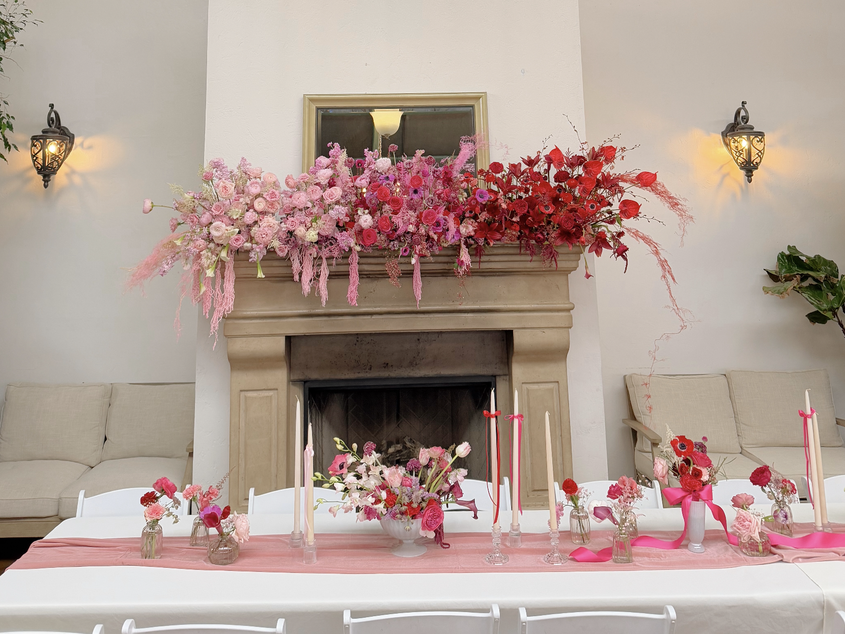 A decorated indoor space with a beige fireplace mantel adorned with pink and red floral arrangements. On the table in front, there are pink and red flower centerpieces with candles in glass holders. The background features two beige couches, a mirror