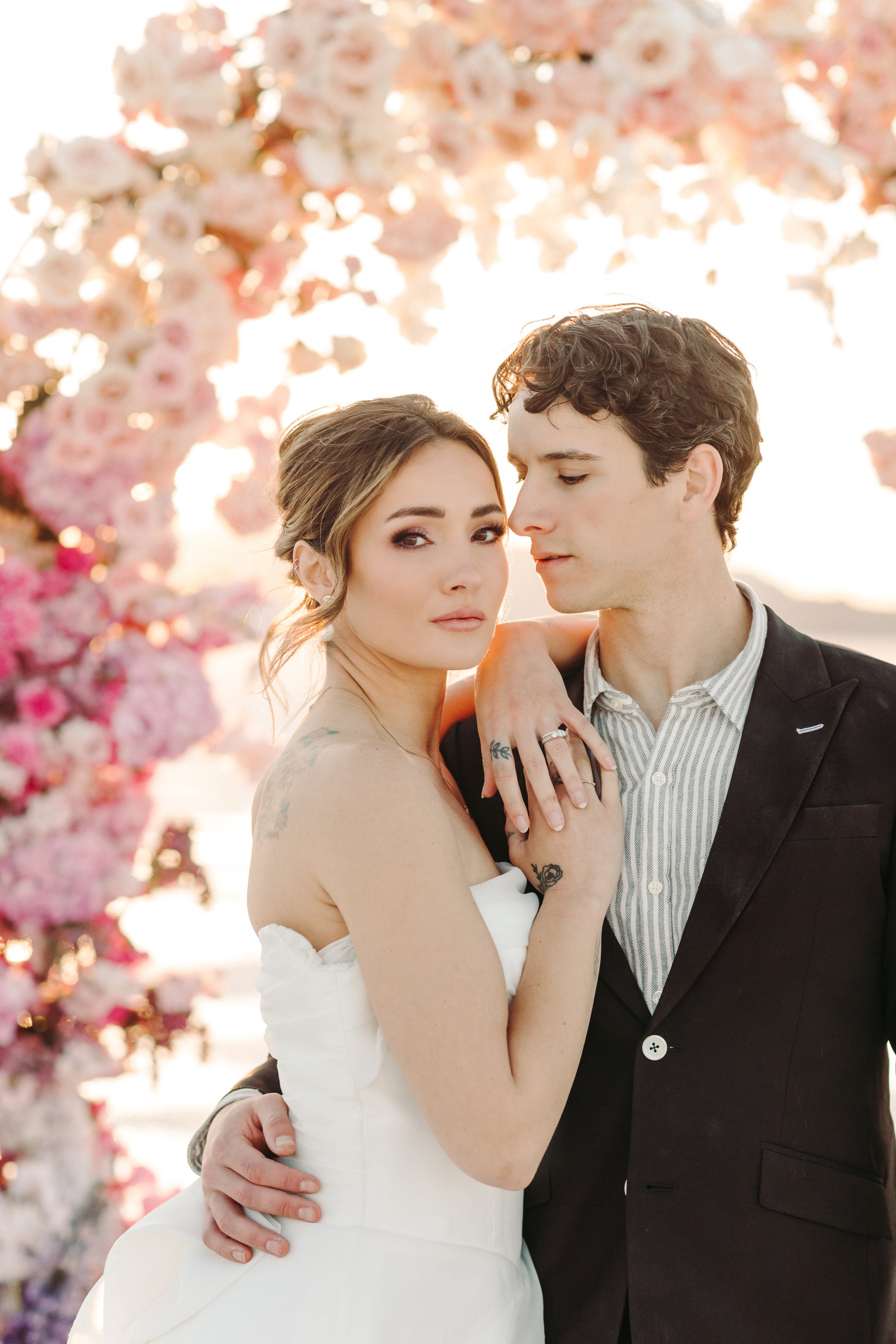 A bride and groom pose closely together outdoors during sunset, with pink flowers in the background. The bride has light brown hair styled in loose waves and is wearing a white wedding dress. The groom has dark curly hair and is wearing a black suit 