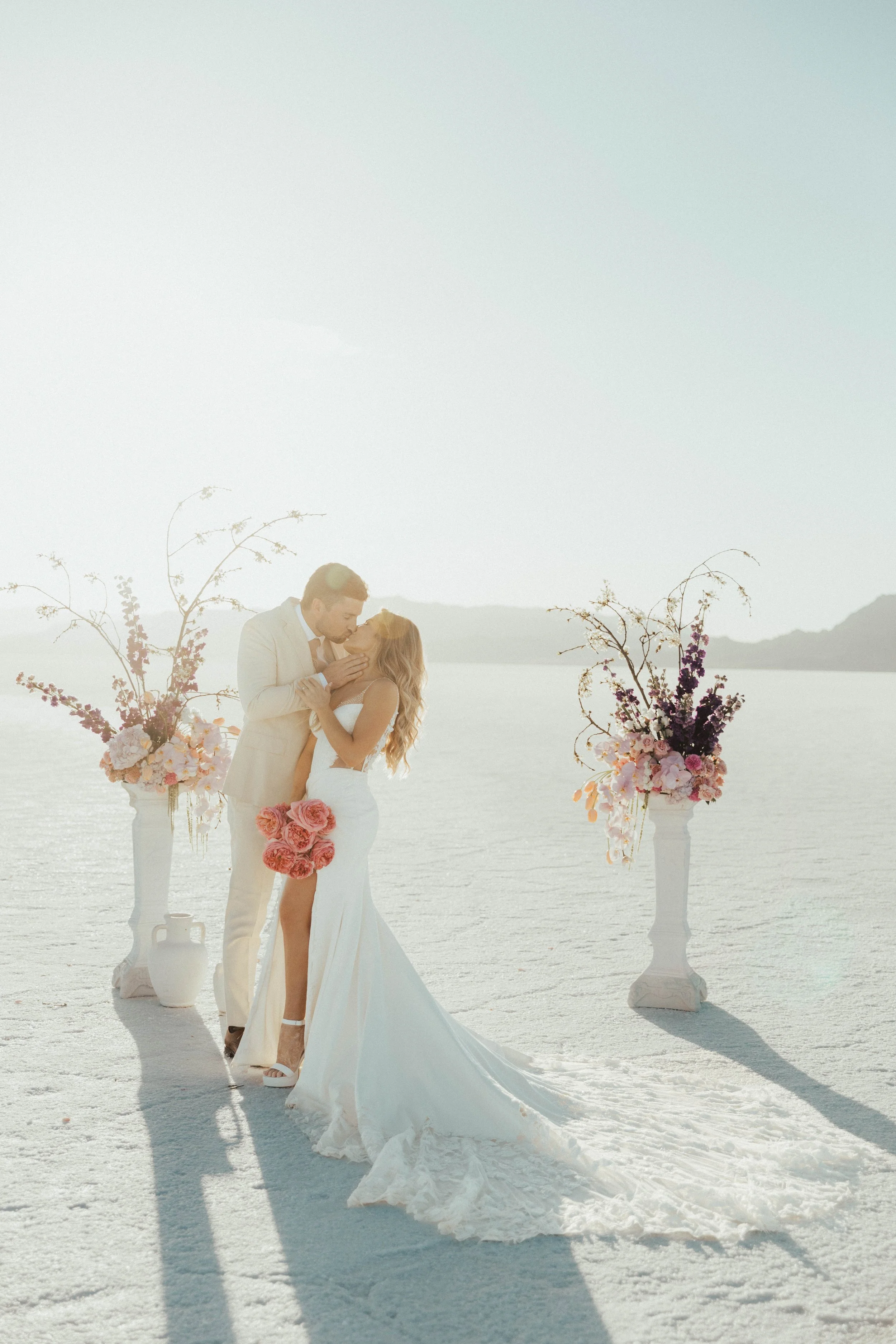 A bride and groom sharing a romantic moment at their wedding ceremony in a desert setting, with decorative floral arrangements and a scenic backdrop of mountains and water.