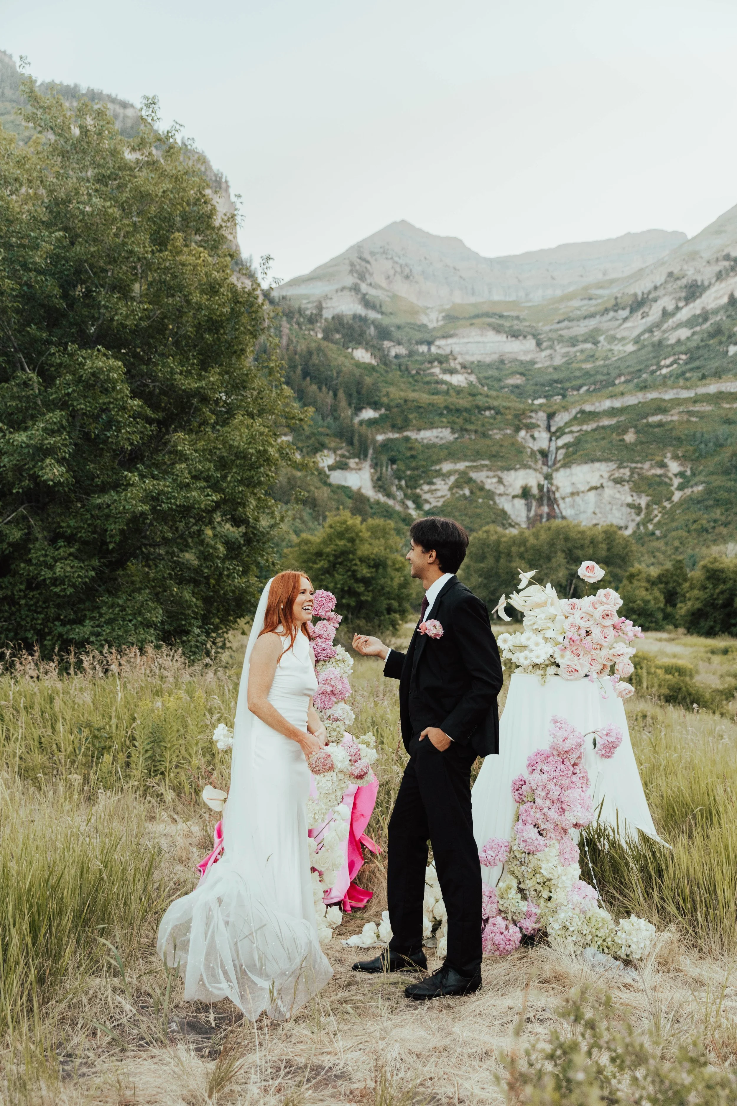 A wedding ceremony outdoors in a grassy field with trees and mountains in the background. A bride with red hair and a white dress holds a bouquet of pink and white flowers, smiling at a groom wearing a black suit. Behind them is a floral arrangement 