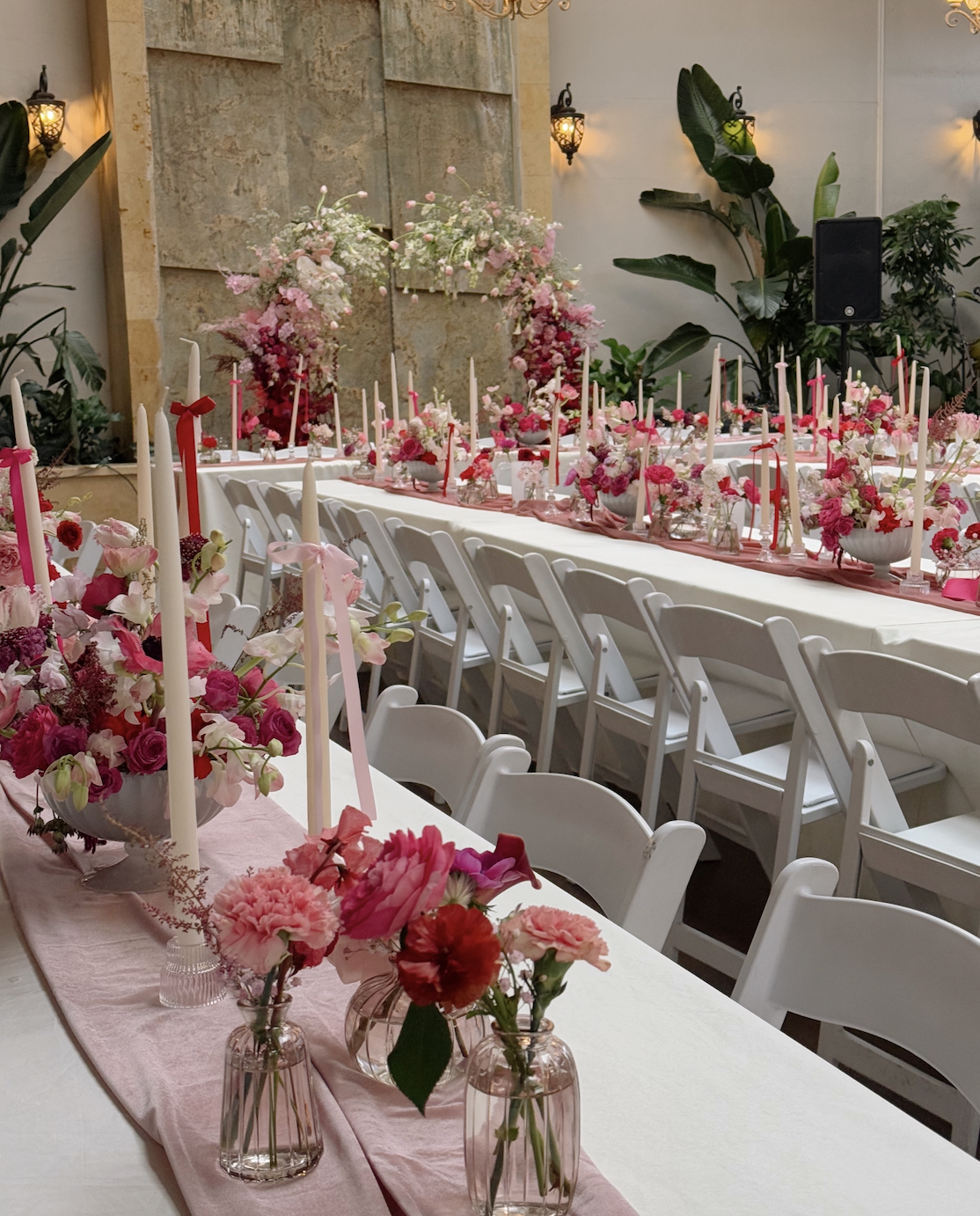 A banquet table decorated with pink and white flowers, candles, and a pink table runner, set for a celebration or wedding in an elegant indoor space with warm lighting and lush green plants.
