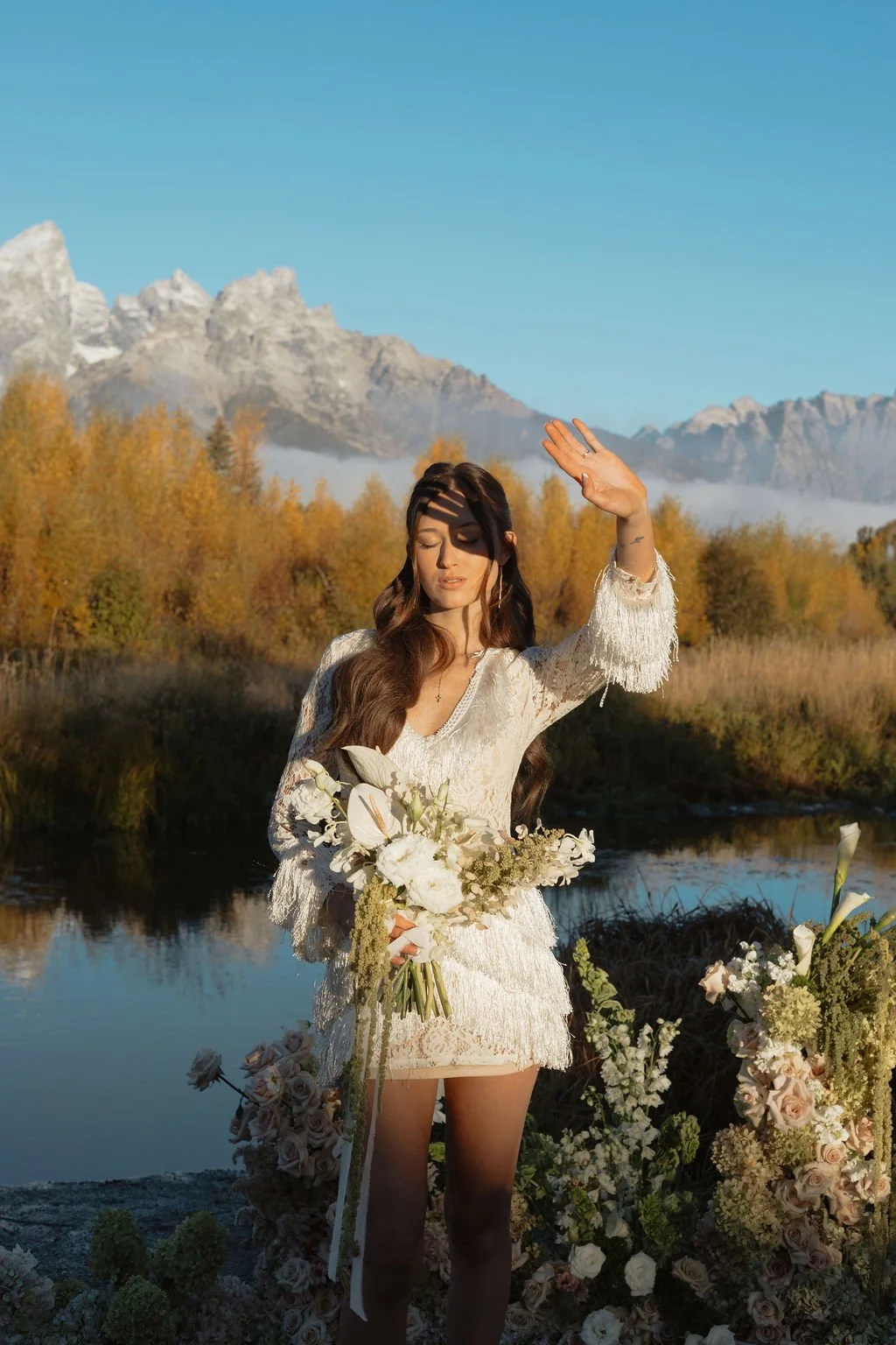 A woman in a white dress holding a bouquet of flowers near a body of water, with mountains and trees in the background on a sunny day.