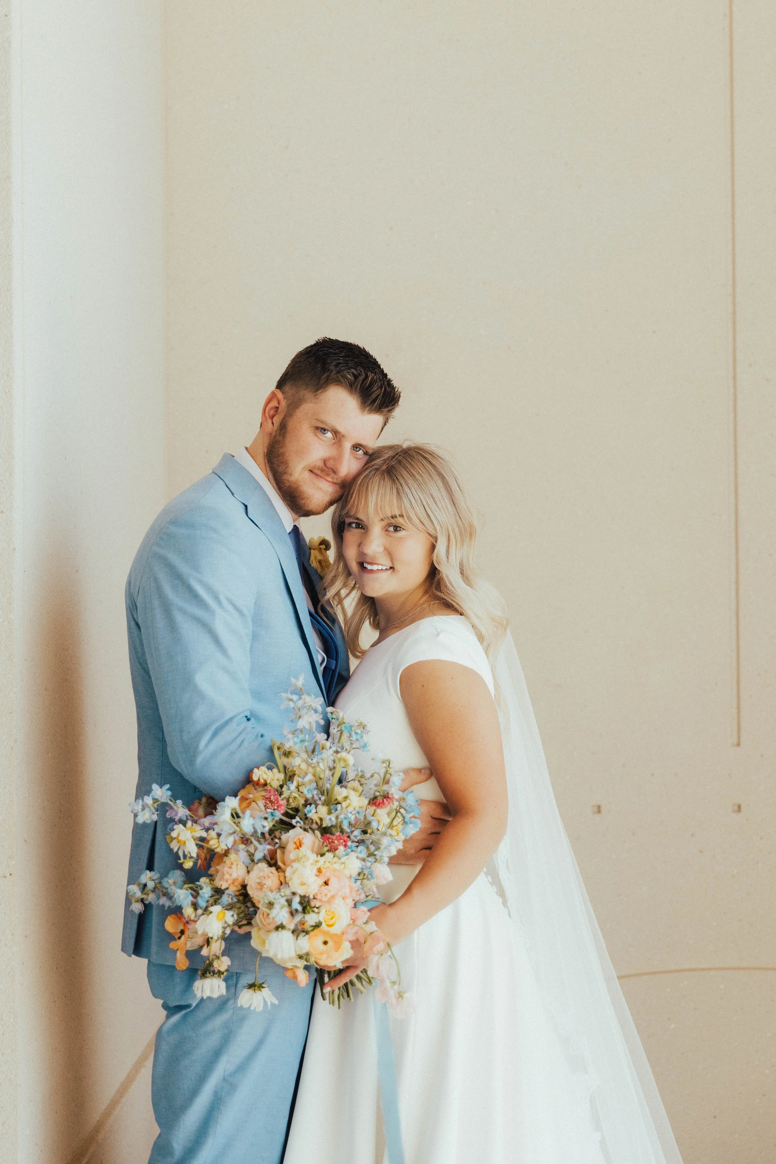 A smiling couple in wedding attire standing against a plain wall, the man in a light blue suit holding a colorful bouquet, and the woman in a wedding dress with blonde hair.