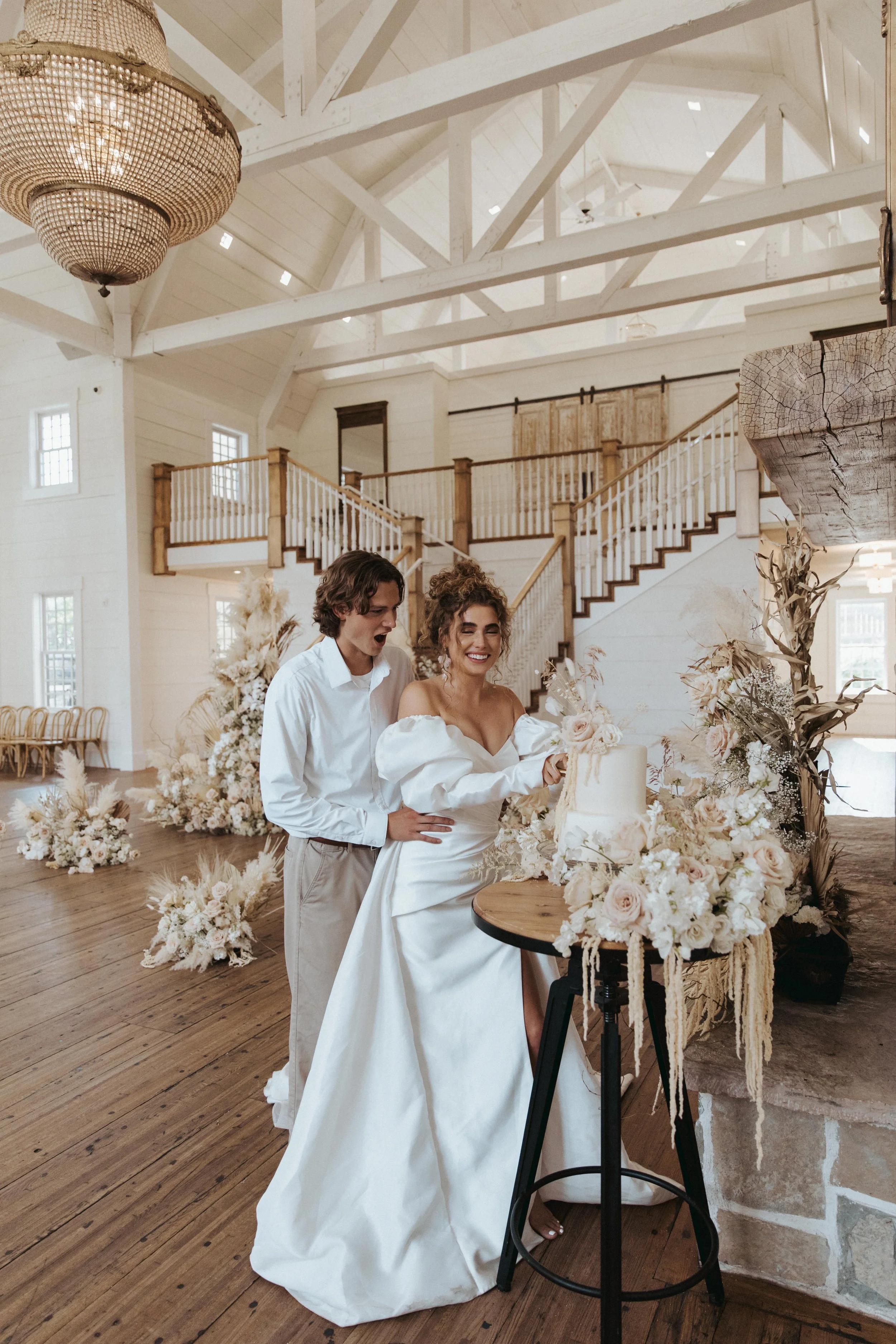 A couple is cutting a wedding cake in a rustic, elegant venue decorated with white flowers and dried plants. The bride is wearing a white wedding dress, and the groom is in a white shirt and beige trousers. The interior features a high ceiling with e