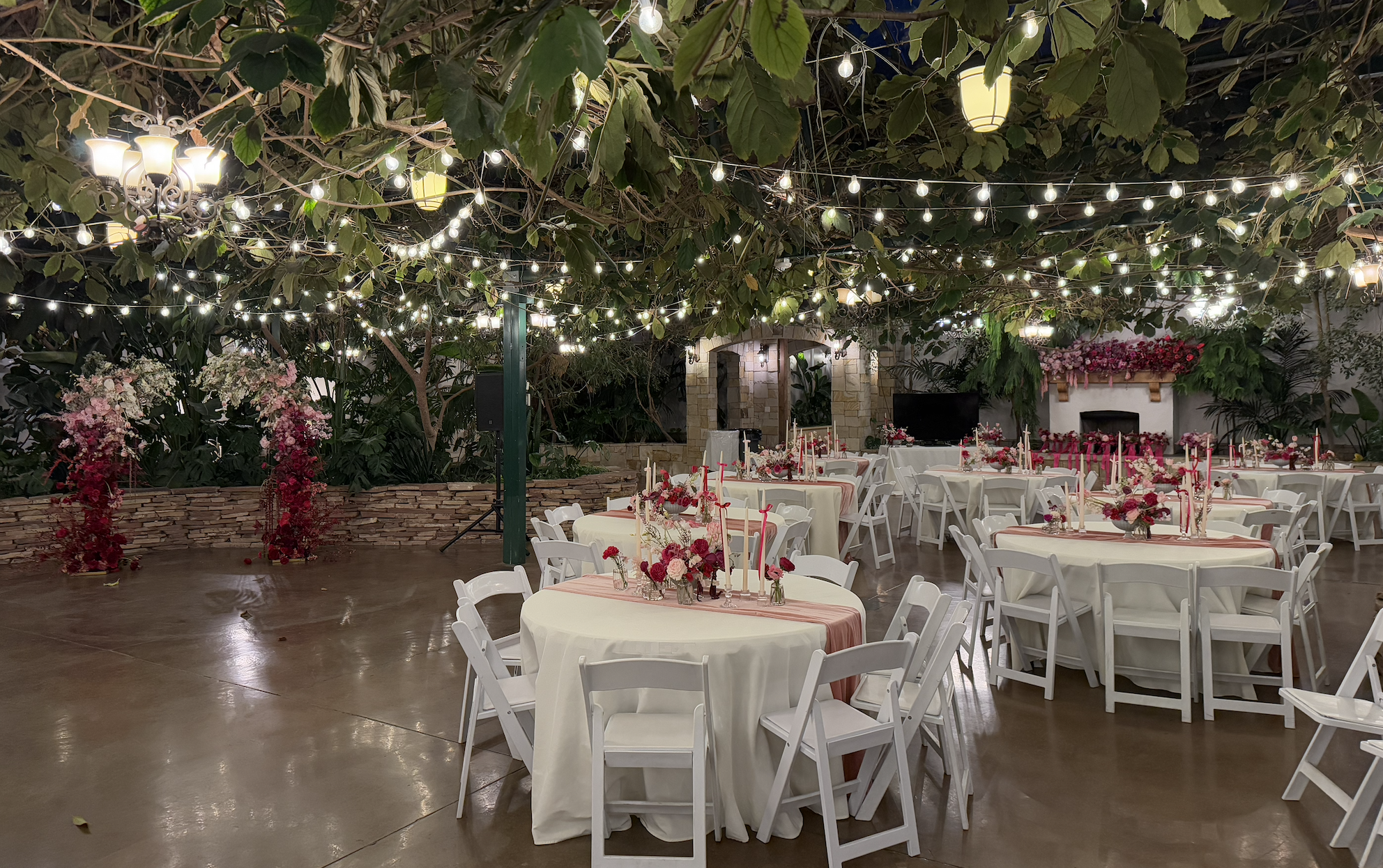 Indoor event space decorated with string lights, hanging lanterns, and floral arrangements, with round tables covered in white tablecloths and pink accents.
