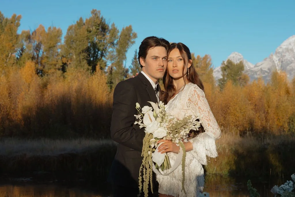 A man in a black suit and a woman in a white lace dress stand close together outdoors, holding a bouquet of white and green flowers, with trees and mountains in the background on a clear day.