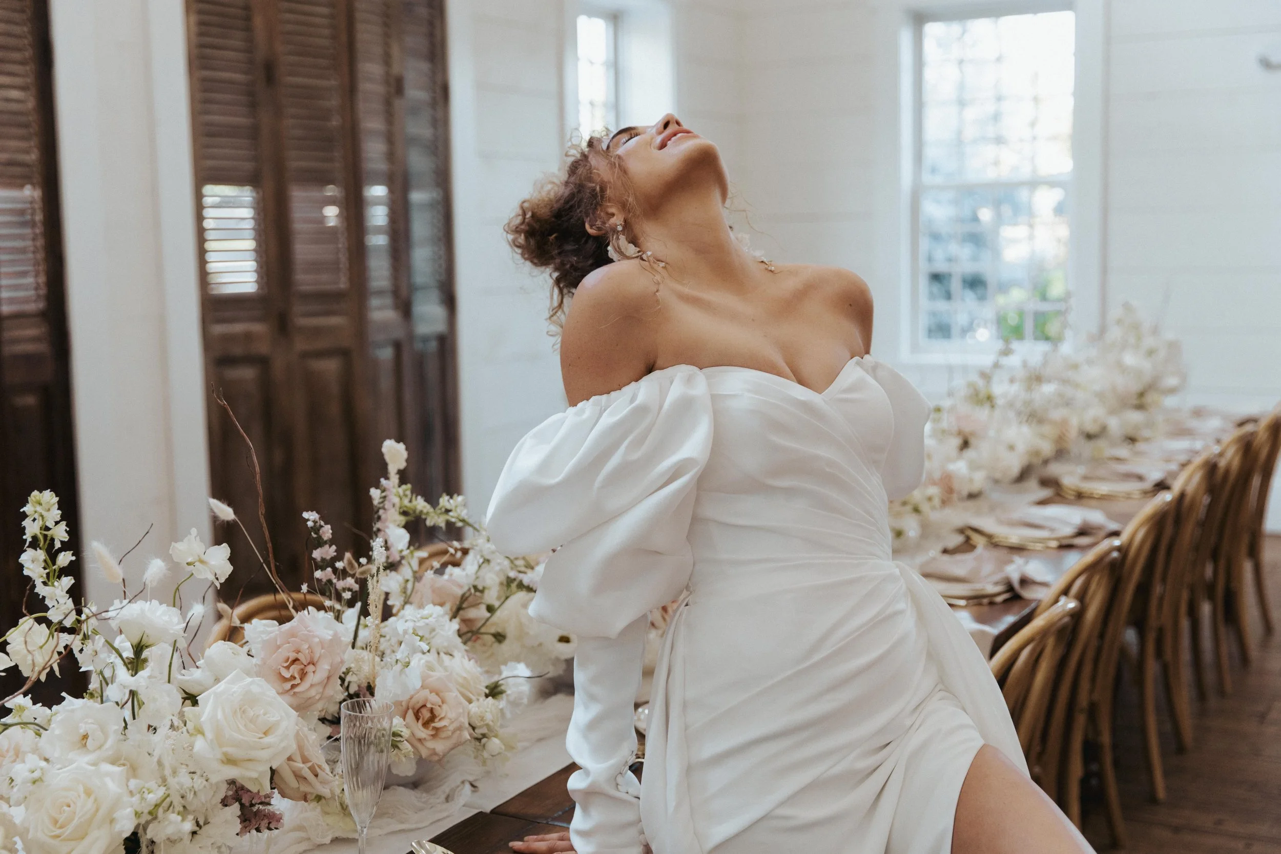 A woman with curly hair in a white dress with puffed sleeves, smiling and leaning her head back, sitting at a table decorated with white and light pink flowers in an airy room with large windows.