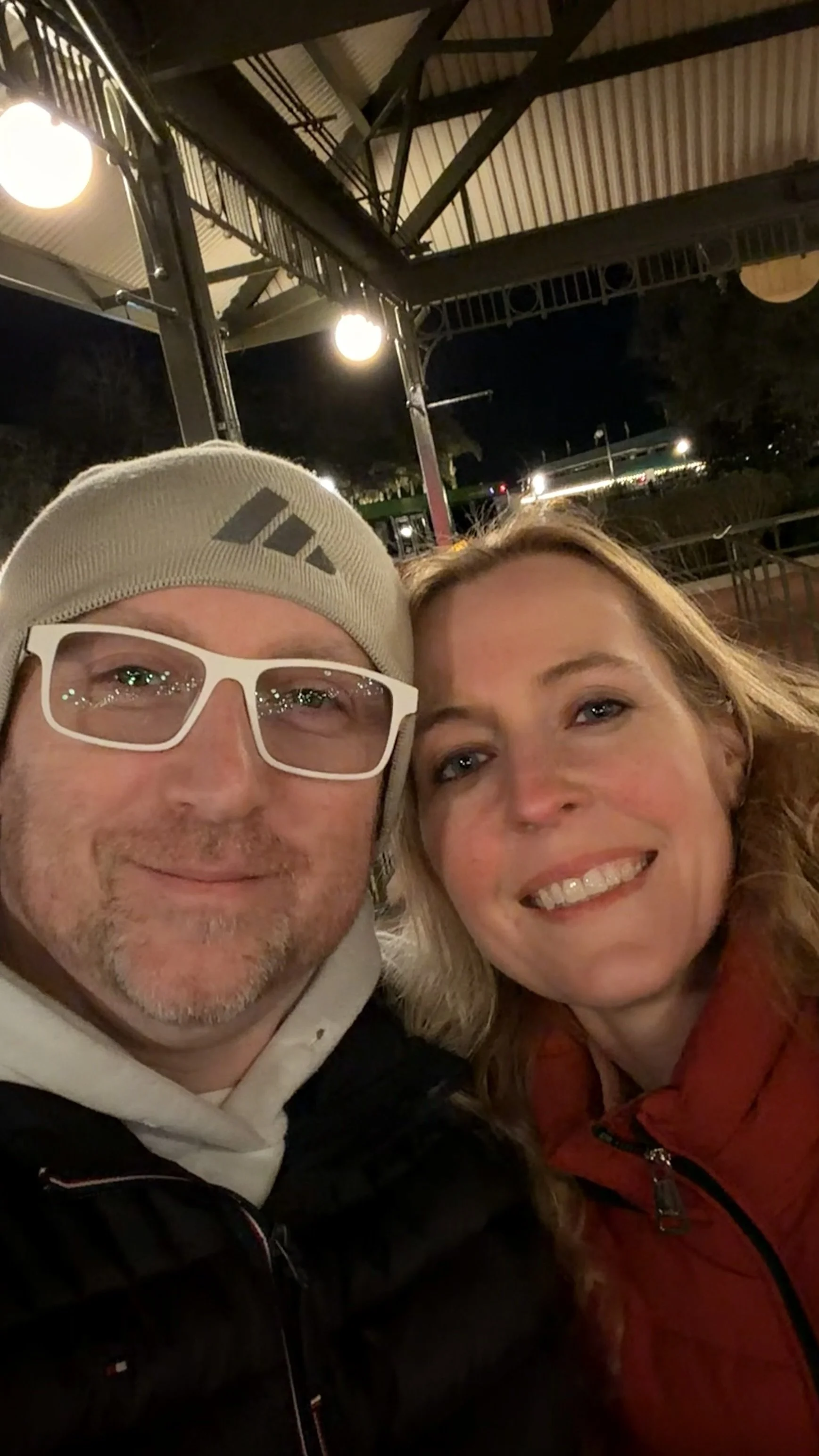 A close-up of a man and woman smiling outdoors at night, with a building's metal roof and lights in the background.