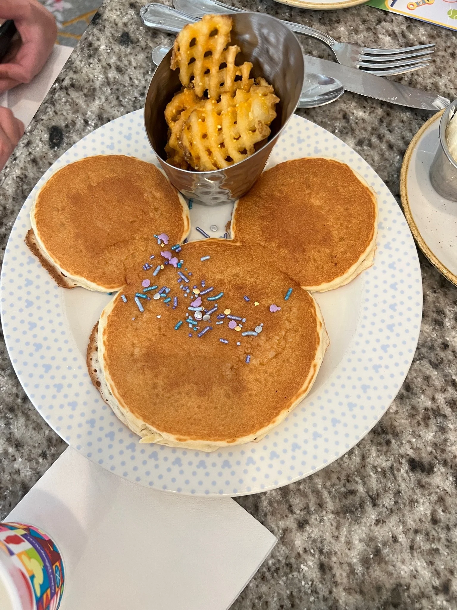 A plate with three pancakes topped with colorful sprinkles, a cup of waffle fries, and a set of silverware on a granite countertop.