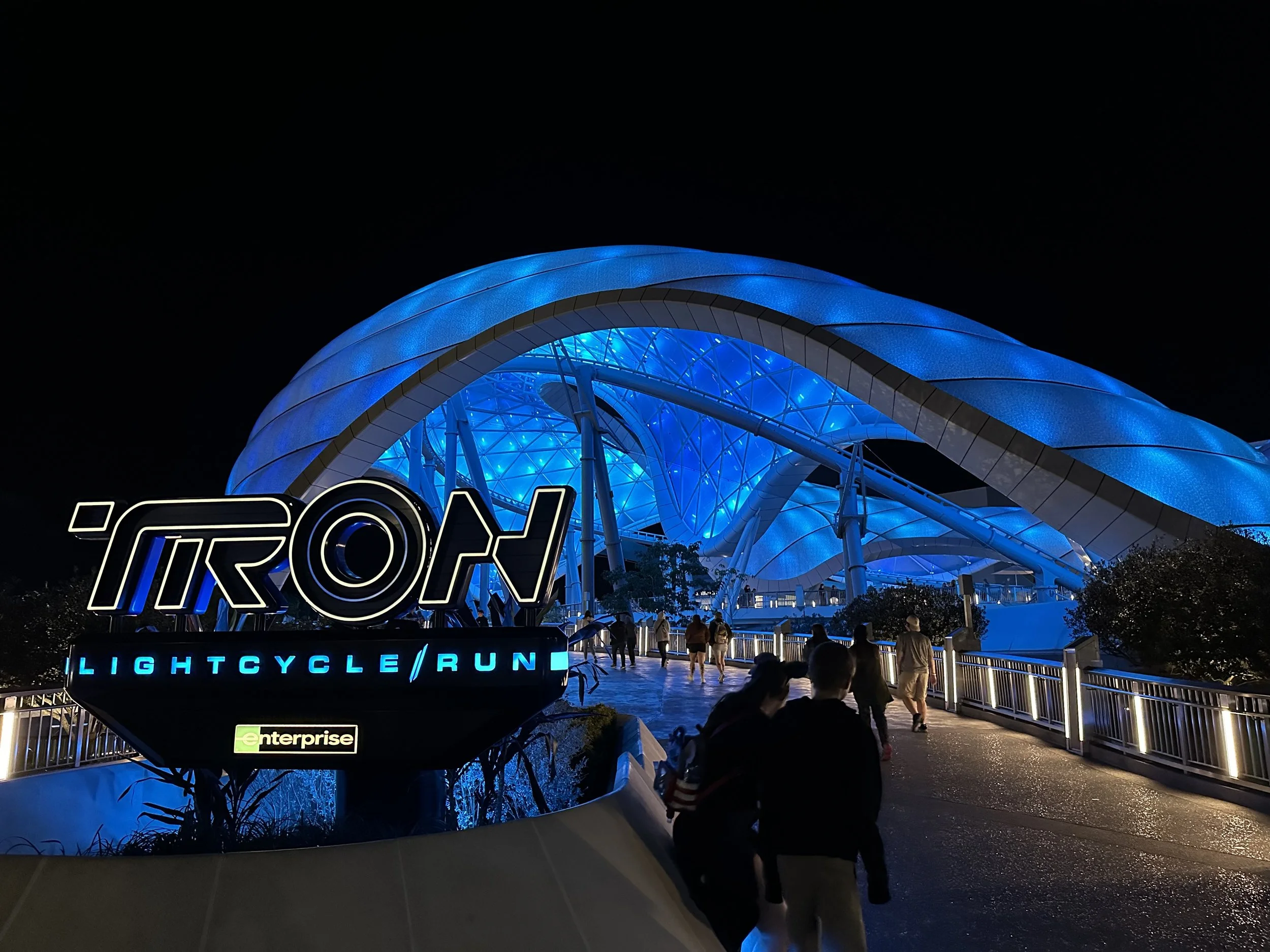 Night view of the futuristic blue glass and metal structure at the entrance of Tron Lightcycle / Run roller coaster at Disney World's Epcot, with people walking towards it.