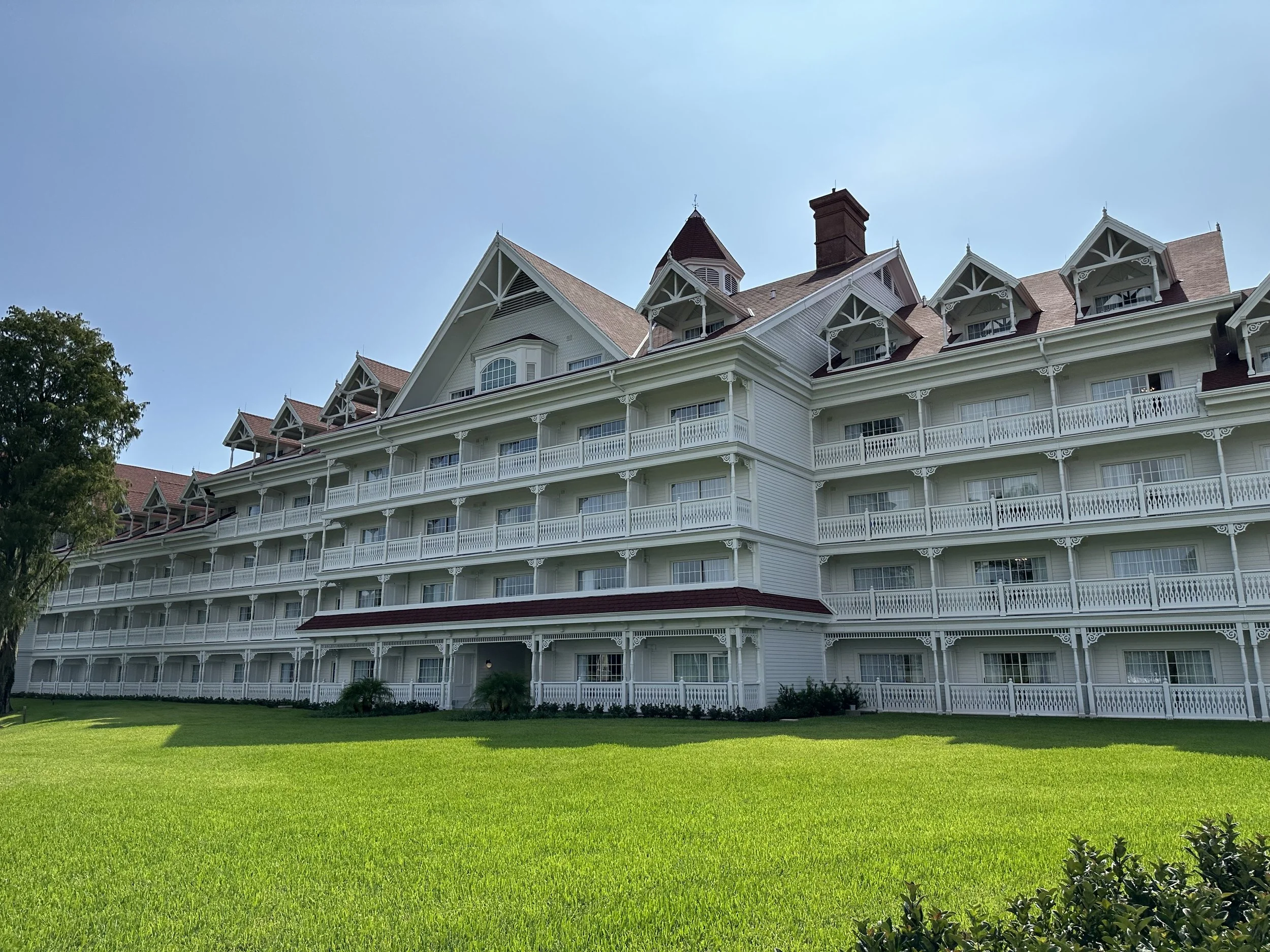 A large, white, multi-story hotel with multiple balconies and a steep, gabled roof. The building is set against a bright blue sky with a well-manicured green lawn in the foreground.