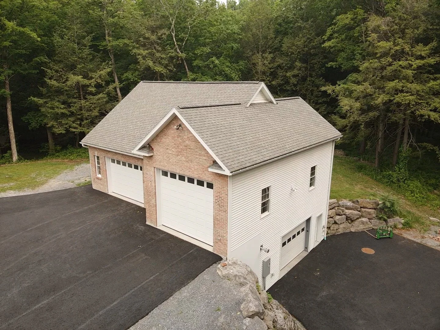 A two-story house with a garage, surrounded by trees, with a paved driveway and a grassy area nearby.