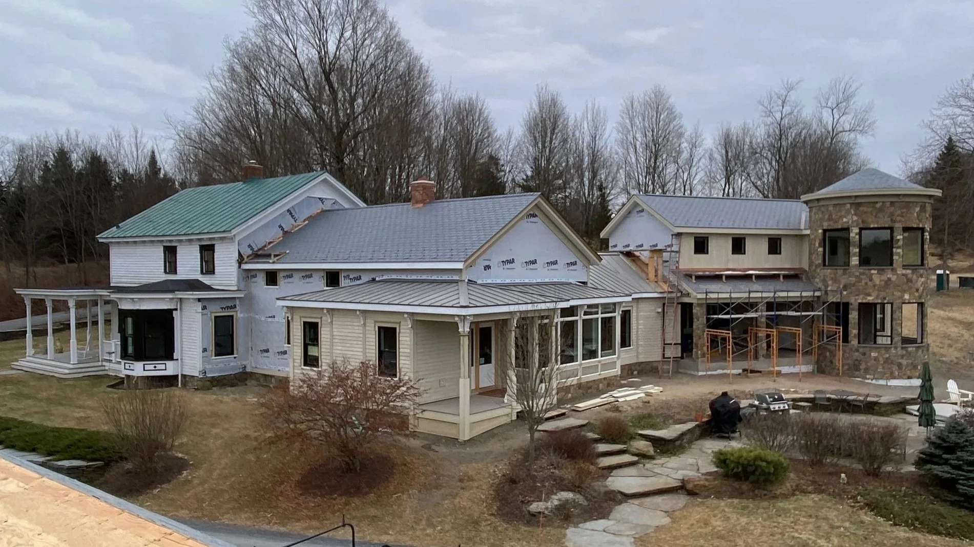 Large house under construction with scaffolding, stone tower, and various roof styles, surrounded by trees and landscaping.
