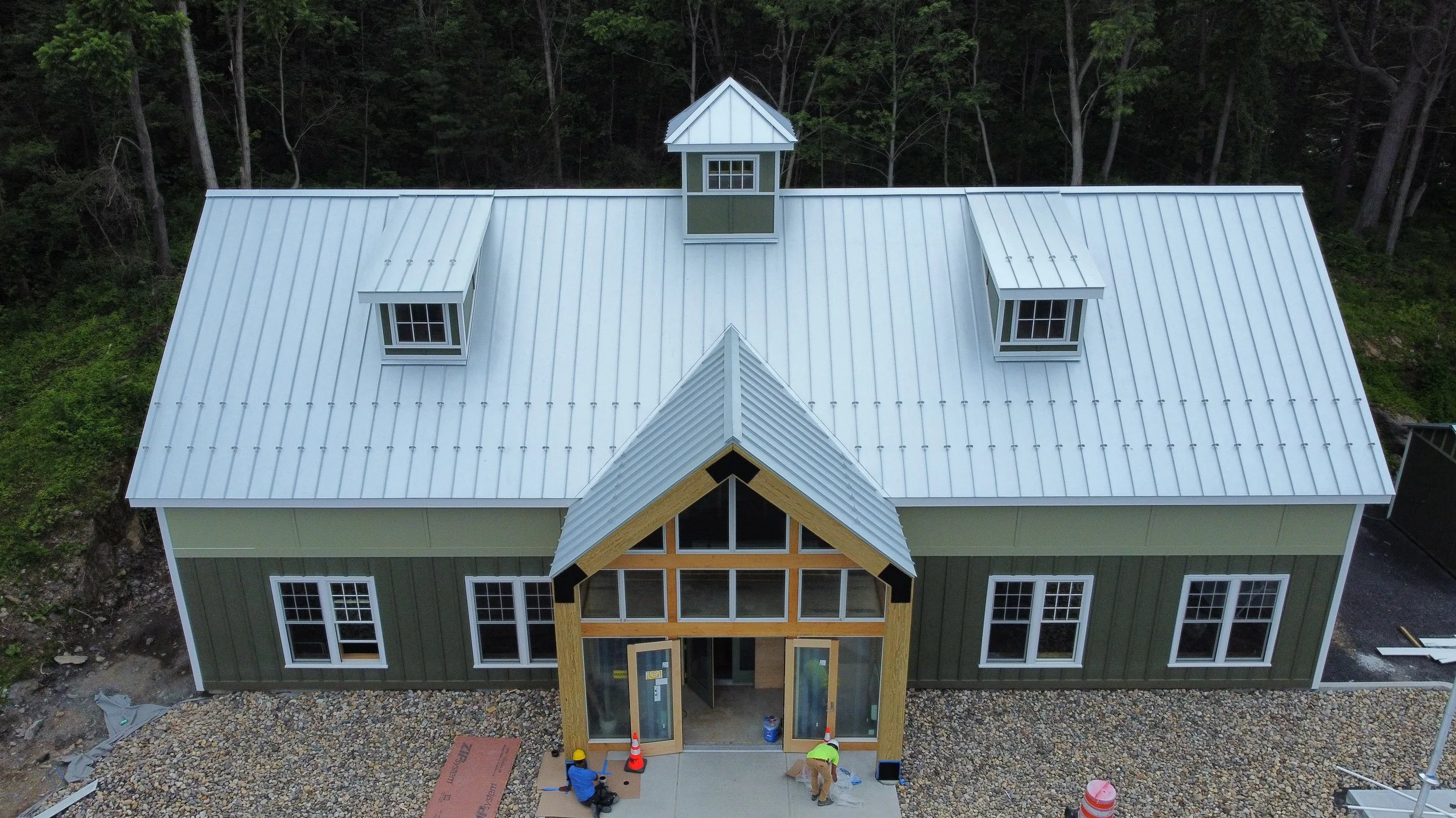 An aerial view of a house under construction with a metal roof, green siding, and a front porch with workers.