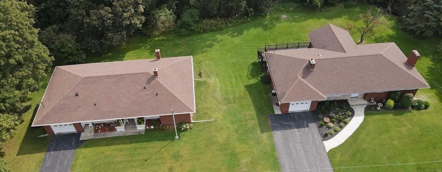 Aerial view of two single-story houses with brown roofs, surrounded by green lawns and trees. The house on the right has a curved walkway and a garden, while the house on the left has a driveway.